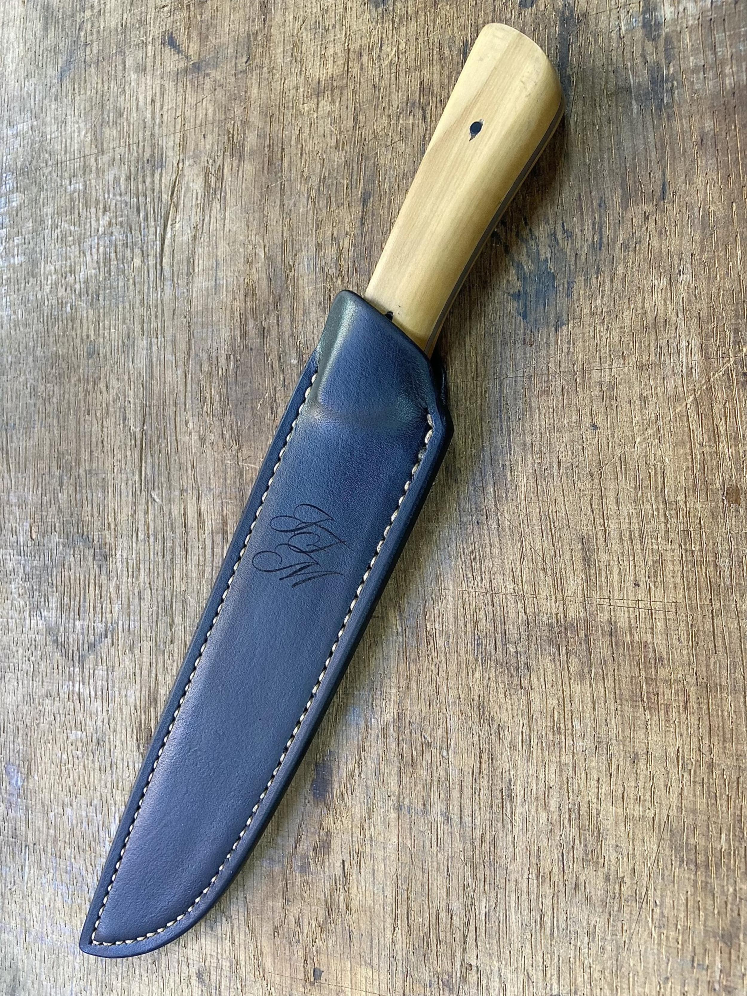 Couteau avec manche en bois clair dans son étui en cuir noir sur fond de bois de chêne.