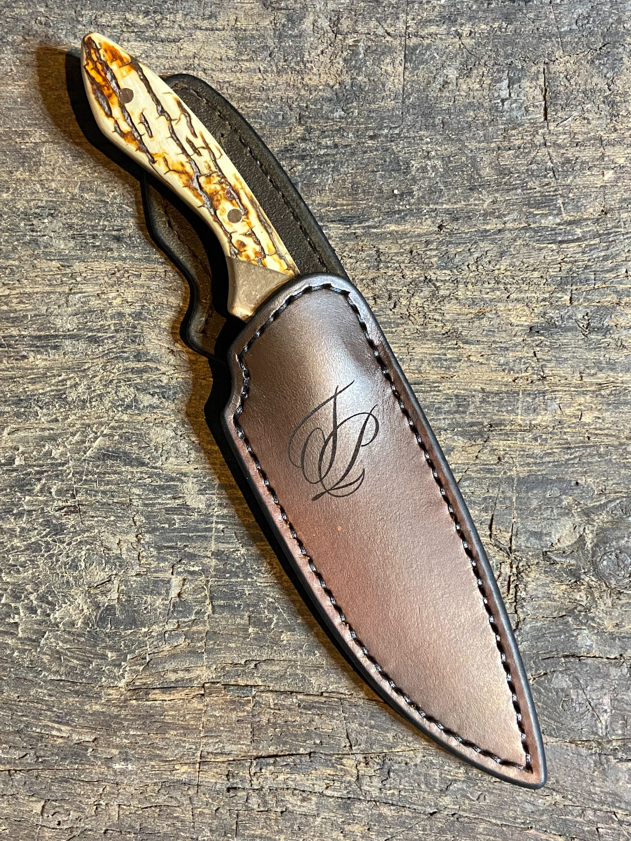 Couteau droit avec manche en défense de mammouth et mitres en bronze massif dans son étui en cuir brun sur fond de bois de chêne.