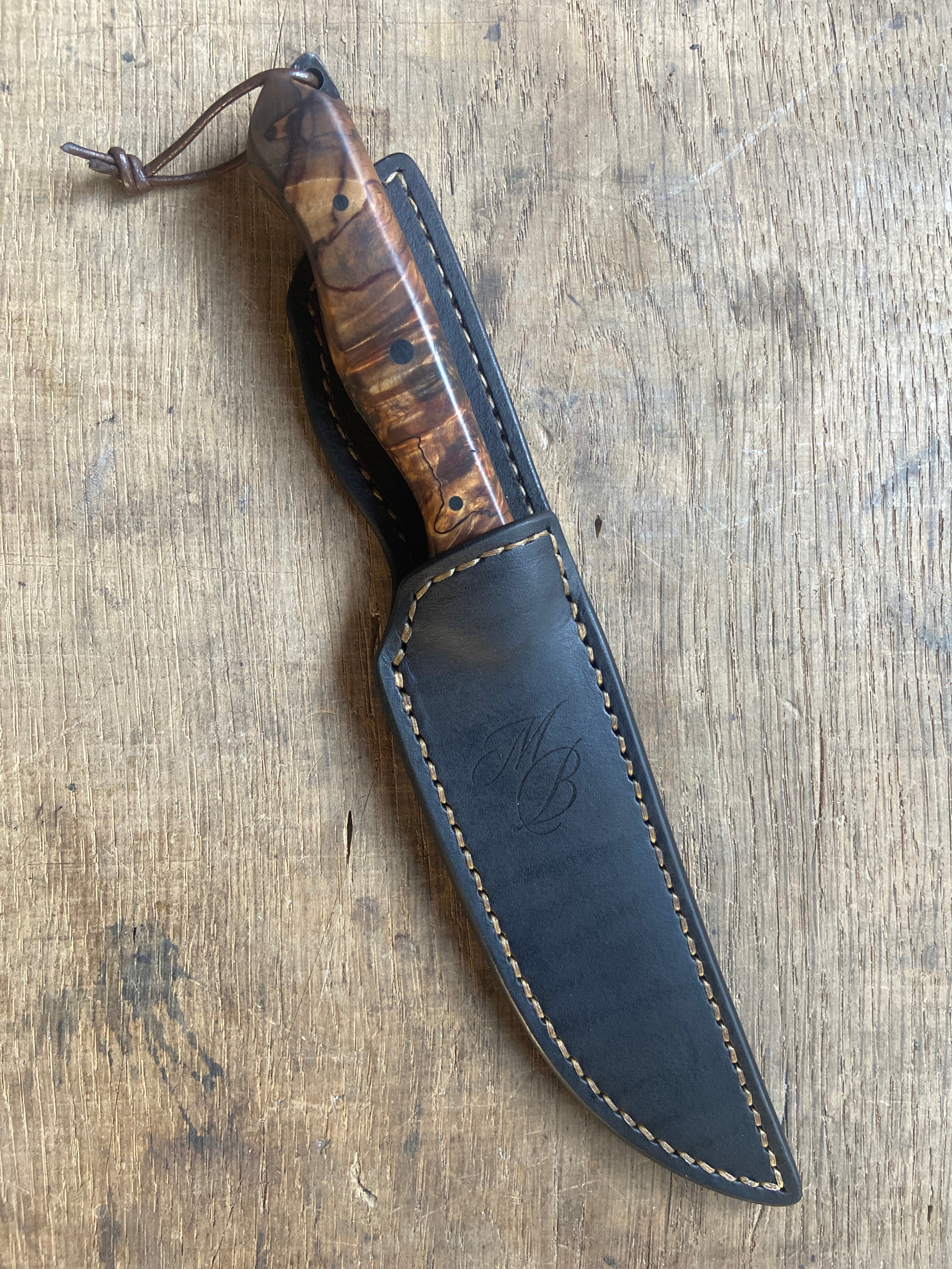 Couteau droit avec manche en bois marron et un lacet en cuir dans son étui en cuir foncé sur fond clair.