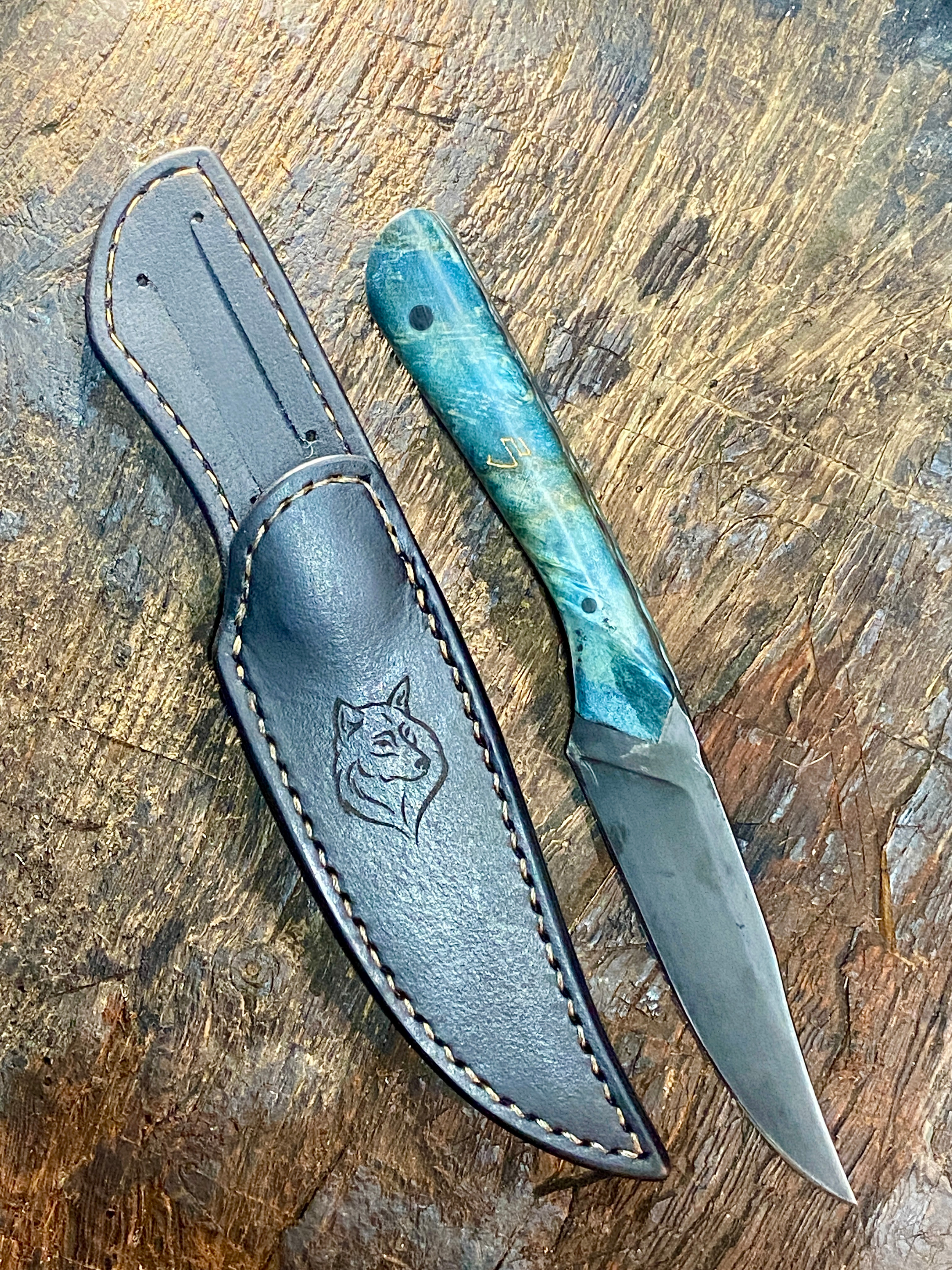 Couteau droit forgé avec manche bleu à côté de son étui en cuir foncé sur fond de bois marron.