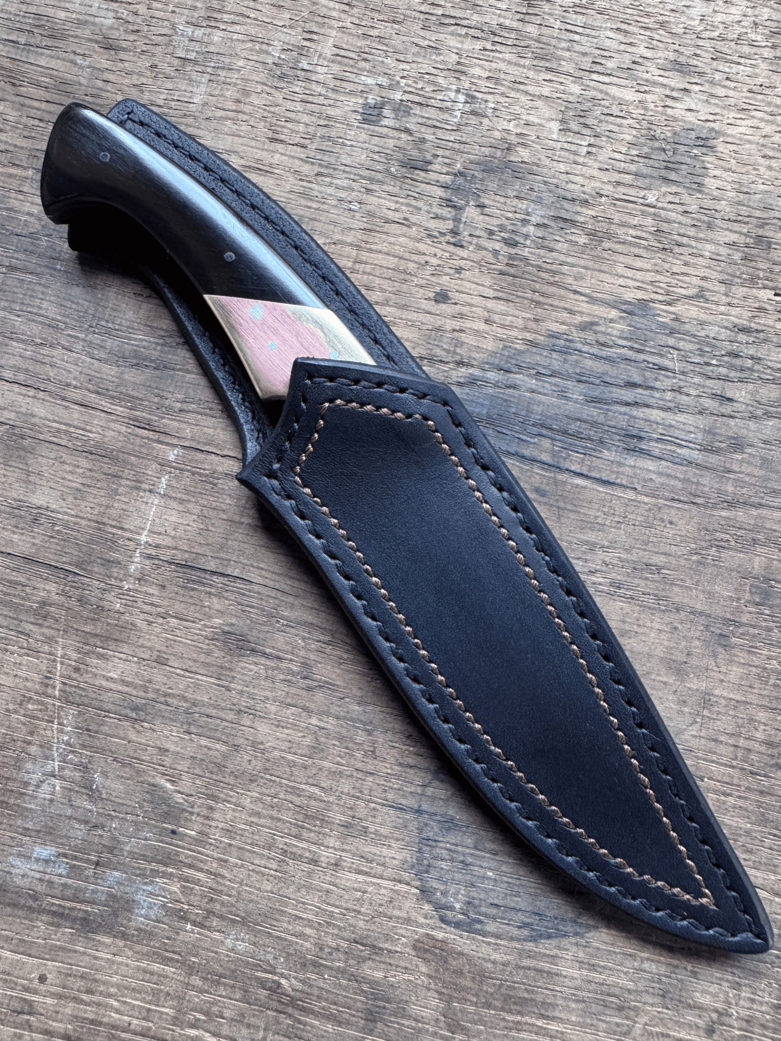 Un couteau avec manche noir dans son étui cuir noir sur fond de bois brun.