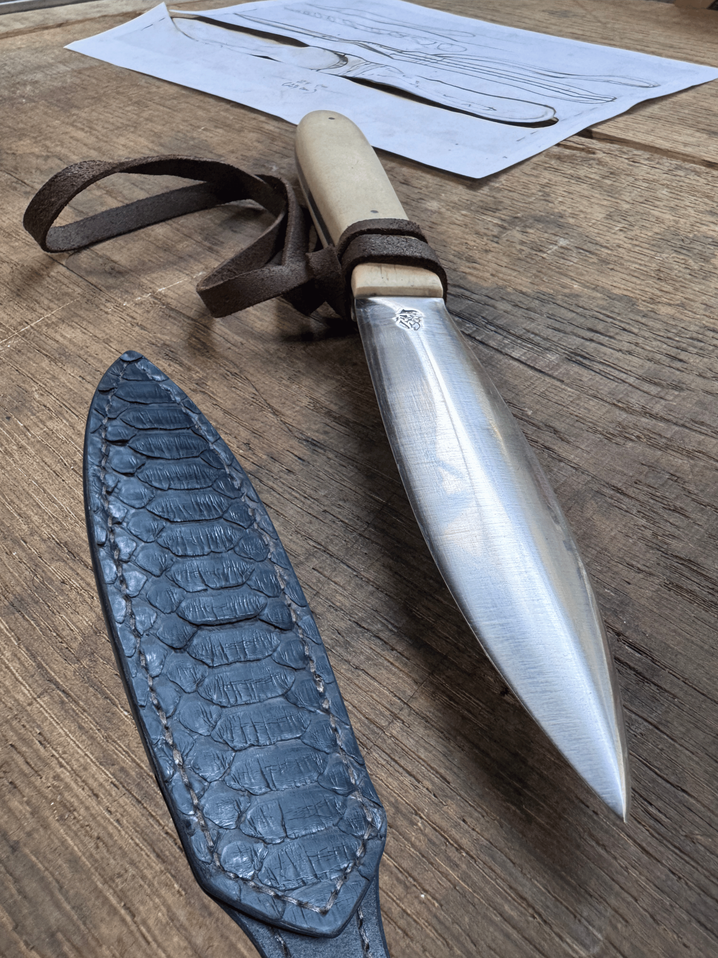Un couteau en acier avec manche blanc et lanière de cuir à côté de son étui de cuir de serpent brun et d'un plan en papier, sur un fond de bois brun.