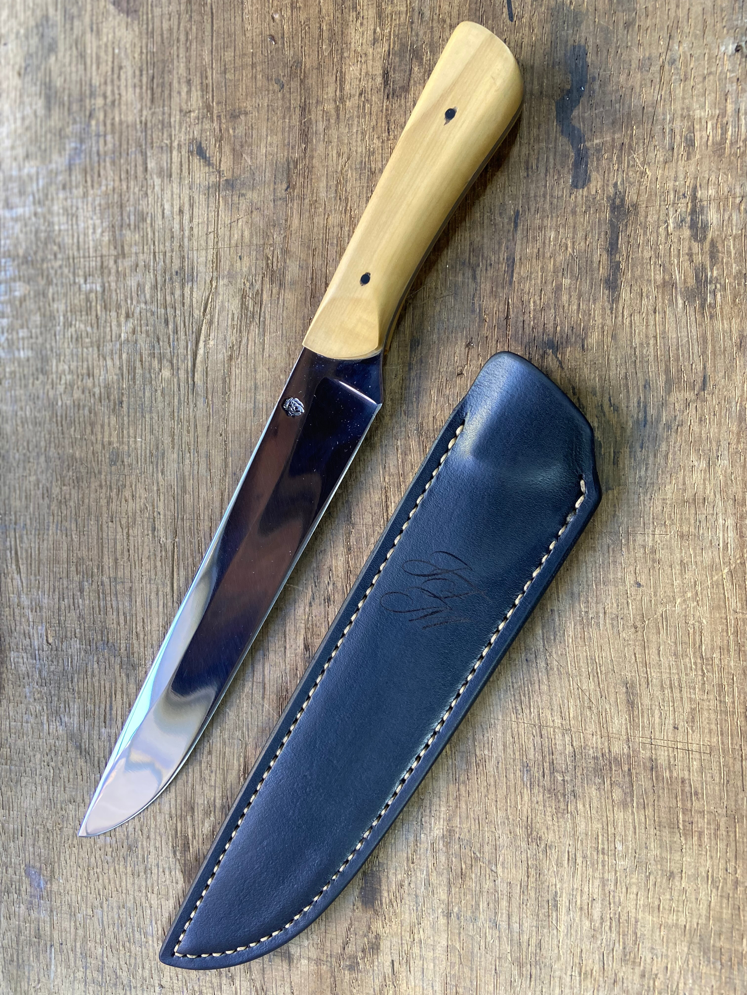 Couteau en acier brillant avec manche en bois clair à côté de son étui en cuir noir sur fond de bois de chêne.