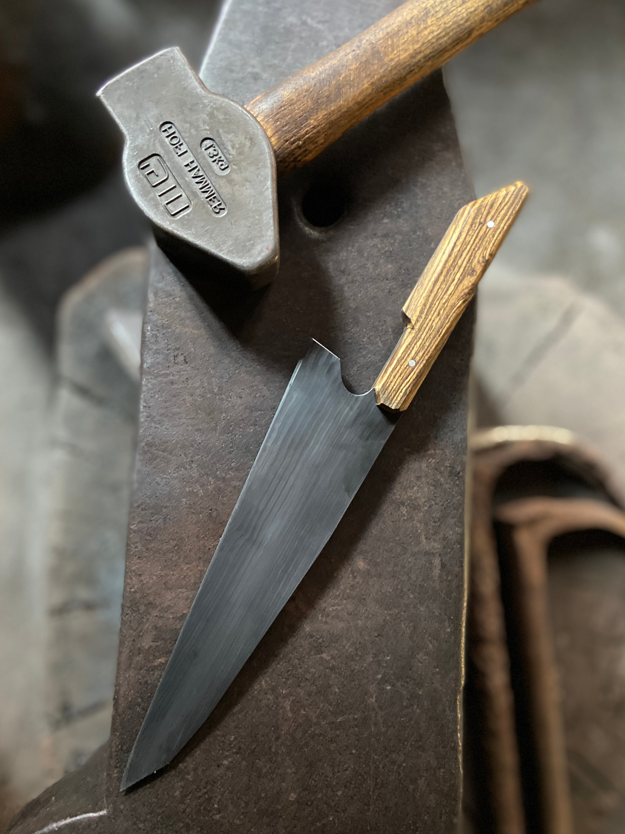 Couteau de Chef damassé avec manche en bois clair à côté d'un marteau de forgeron sur fond d'enclume grise.