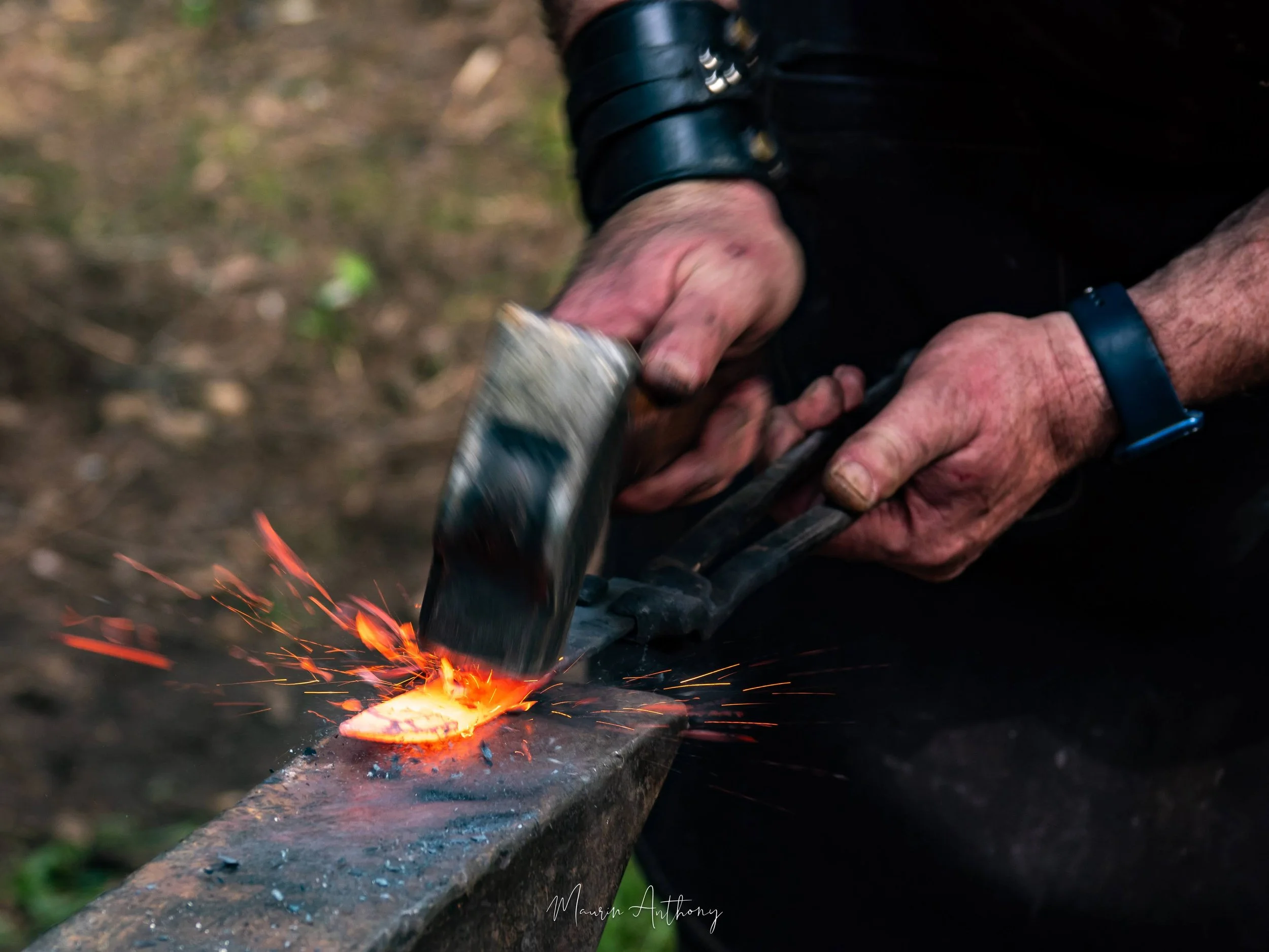 Mains d'un forgeron frappant au marteau une pièce d'acier incandescente sur l'enclume, dans une gerbe d'étincelles