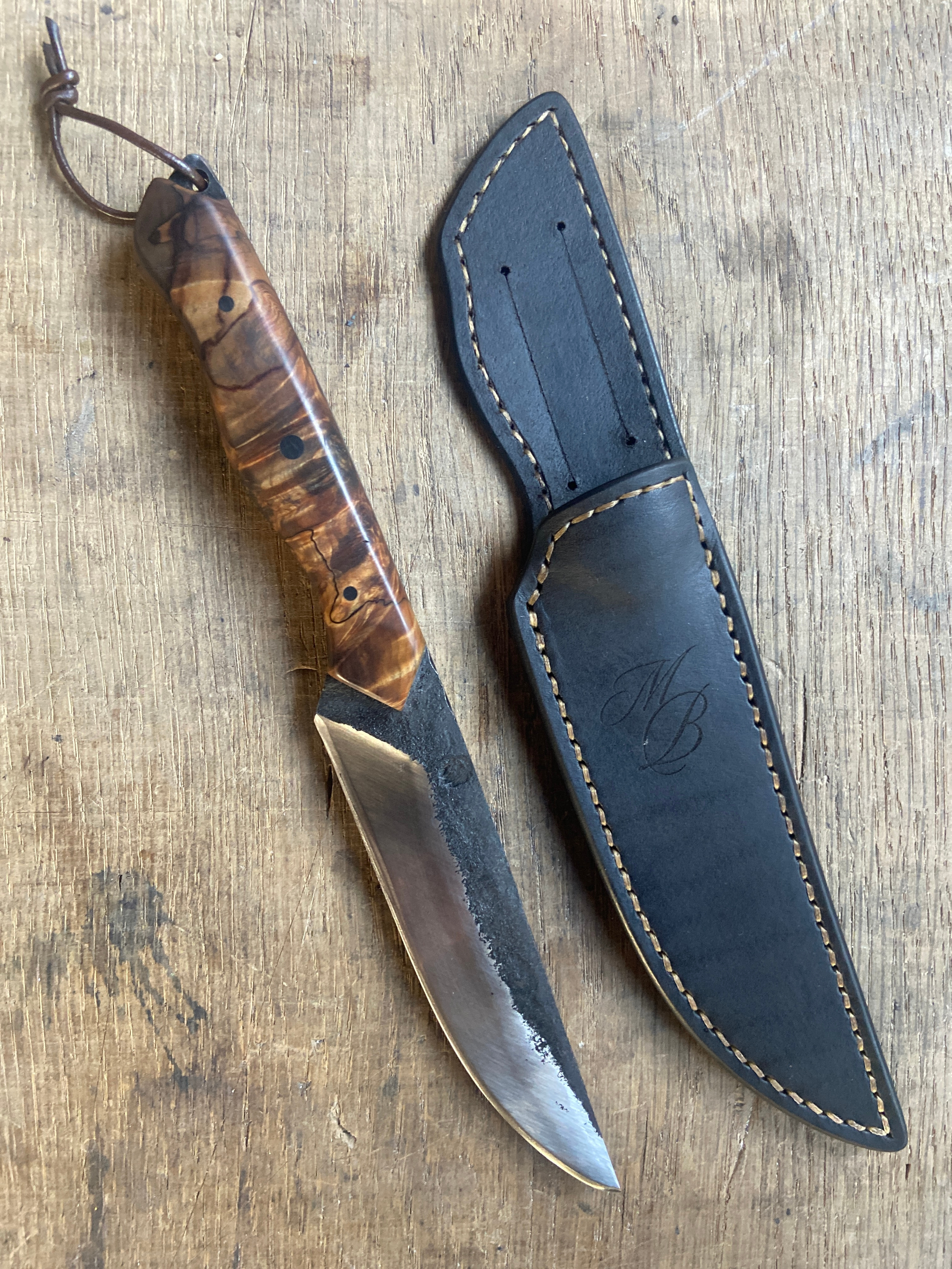 Couteau droit forgé brut de forge avec manche en bois marron et un lacet en cuir à côté de son étui en cuir sur fond de bois clair.