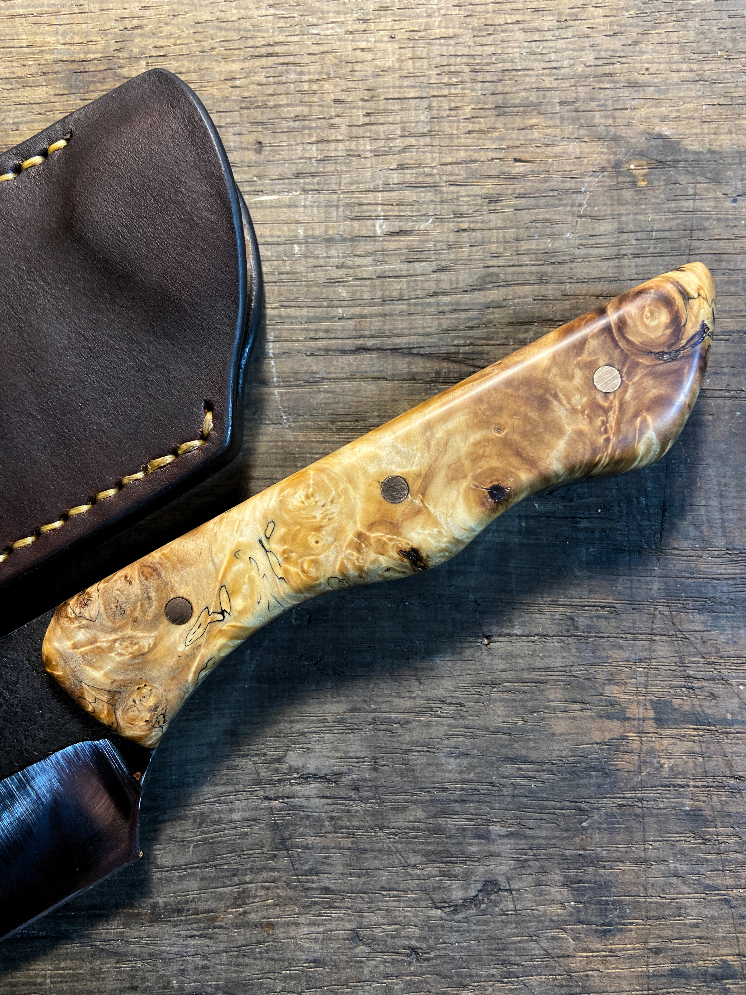 Manche en bois clair sur couteau droit forgé avec étui en cuir sur fond de bois de chêne.