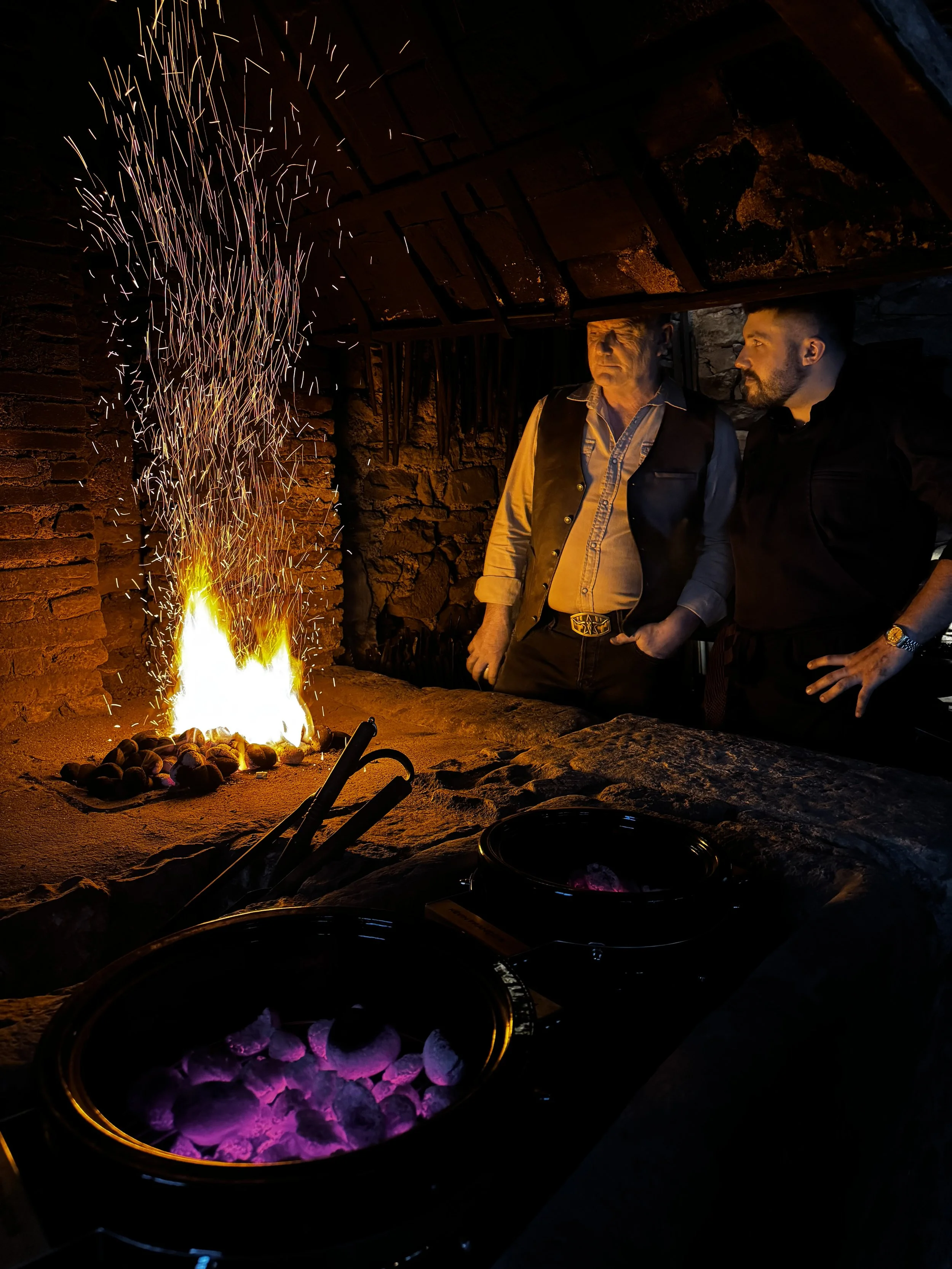 Deux hommes regardent un feu de cheminée dans une pièce en pierre, une des personnes porte une veste en cuir noire et l'autre une veste en tissu avec un col en jean, avec des braises rouges dans le foyer.