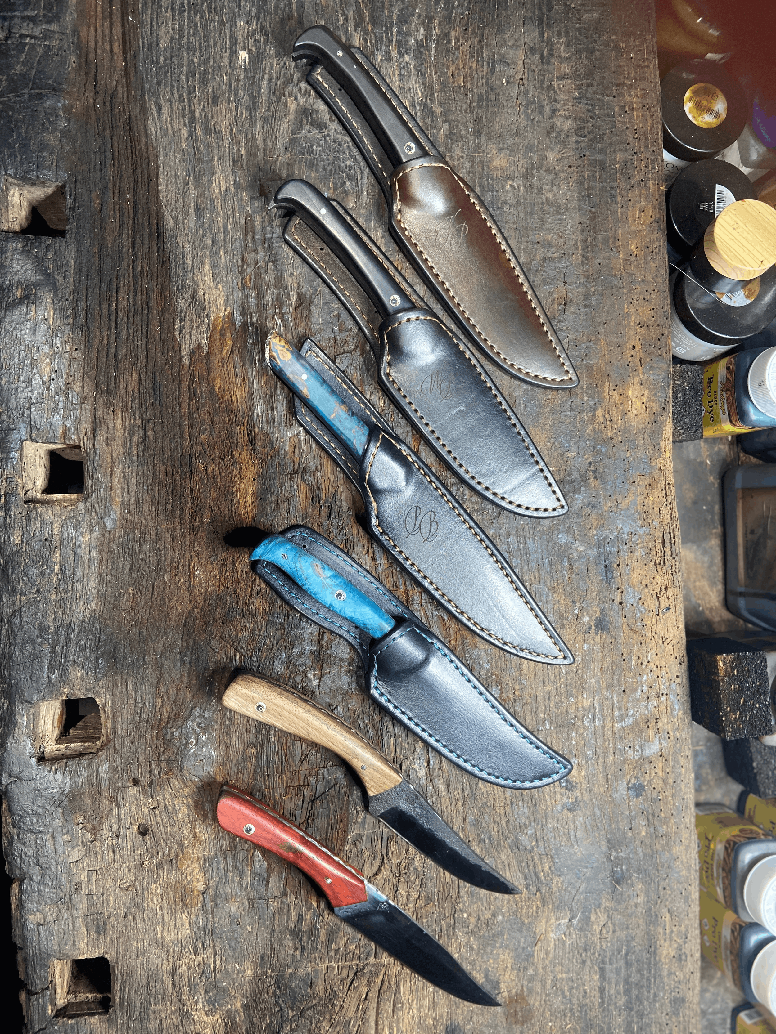 6 couteaux forgés main avec manches colorés et avec leur étui en cuir foncés sur fond de bois foncé.