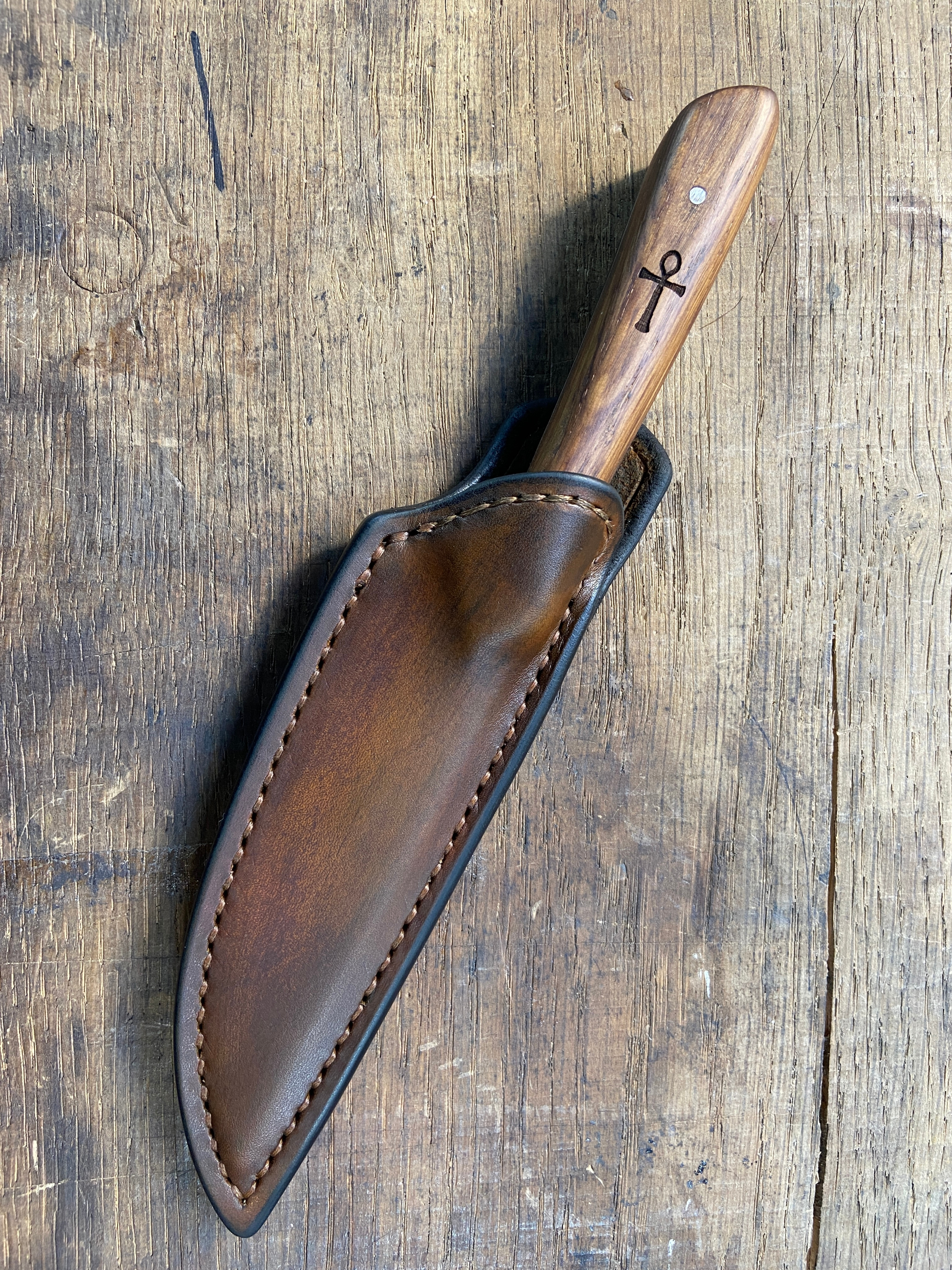 Couteau avec manche en bois brun dans son étui en cuir brun sur fond de bois.