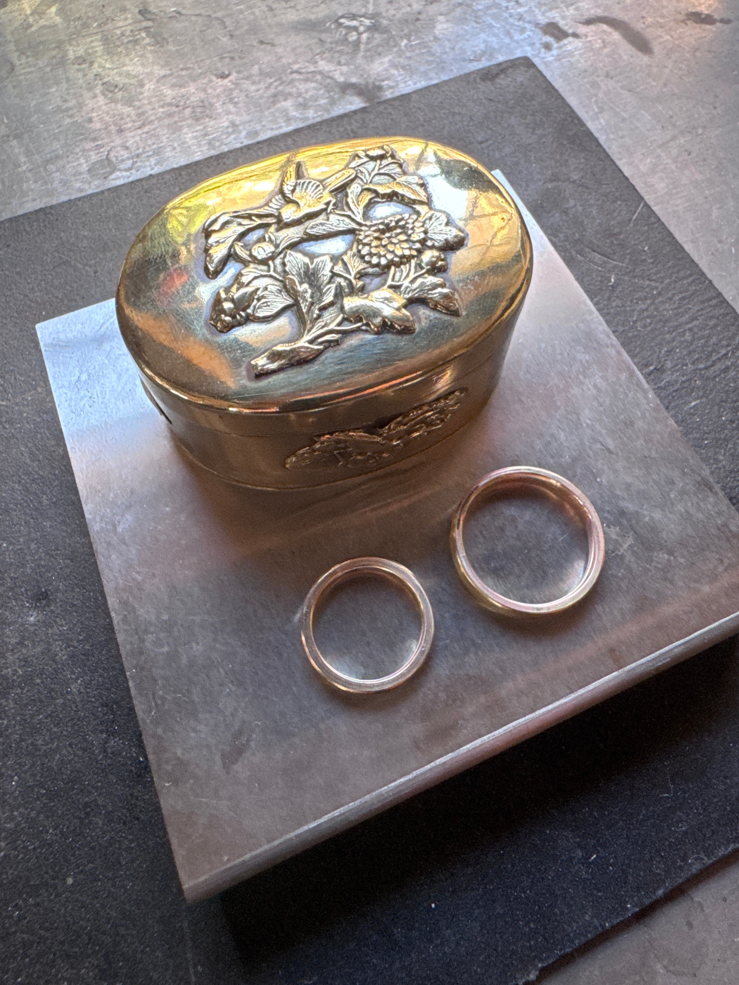 Un coffret en métal doré avec un motif floral relief repose sur une surface en métal. Deux anneaux en métal visibles sont à côté du coffret.