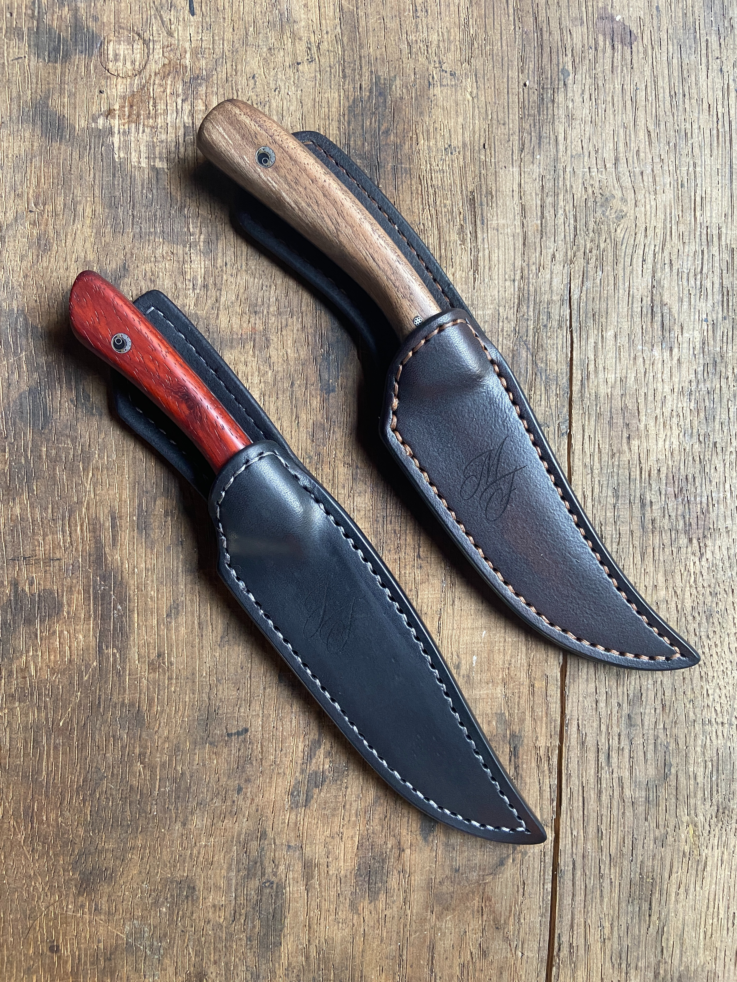 Deux couteaux avec manche en bois rouge et bois clair dans leur étui en cuir foncé sur fond de bois clair.