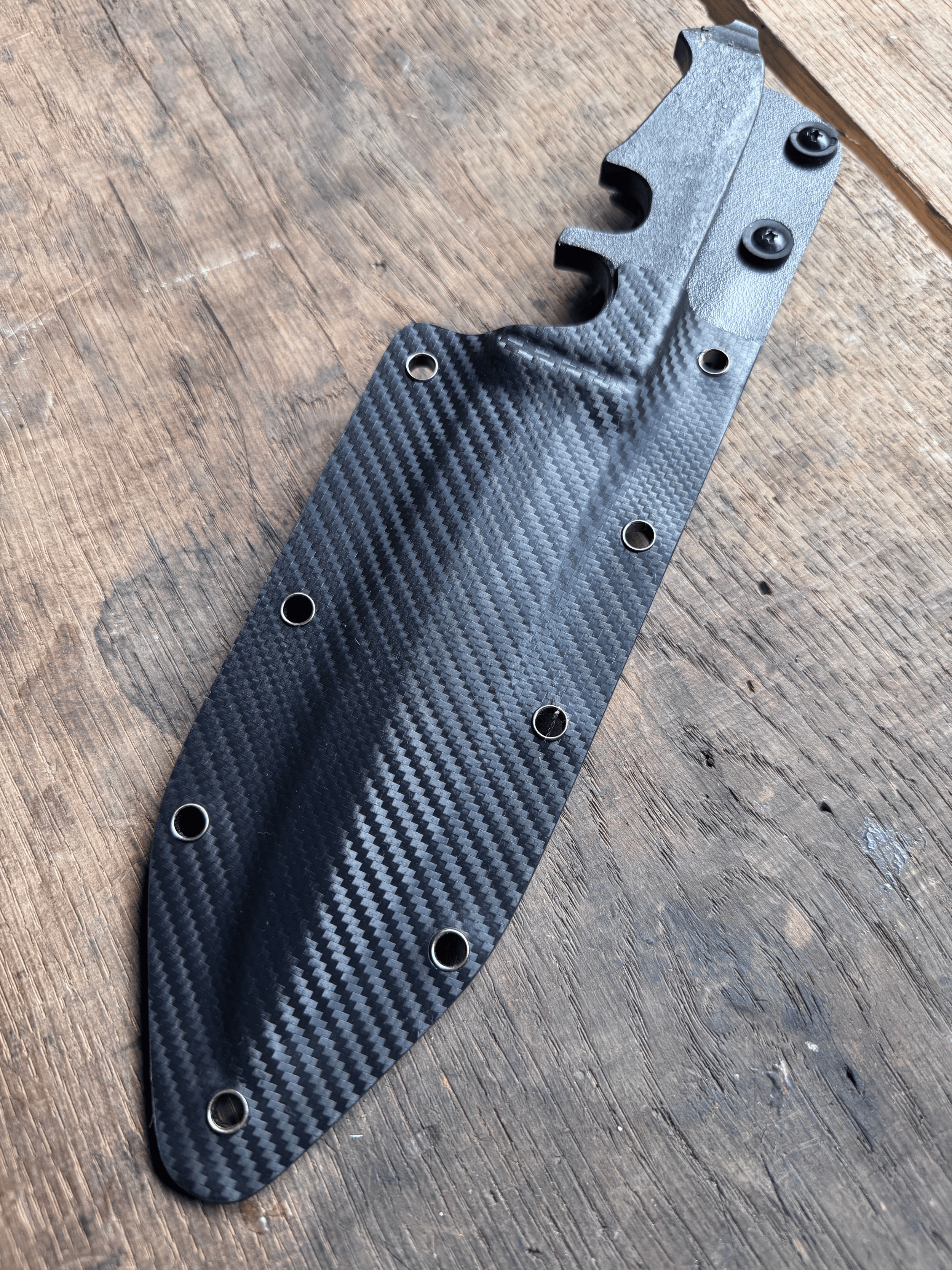 Couteau en acier brut de forge dans un étui Kydex noir sur fond de bois brun.