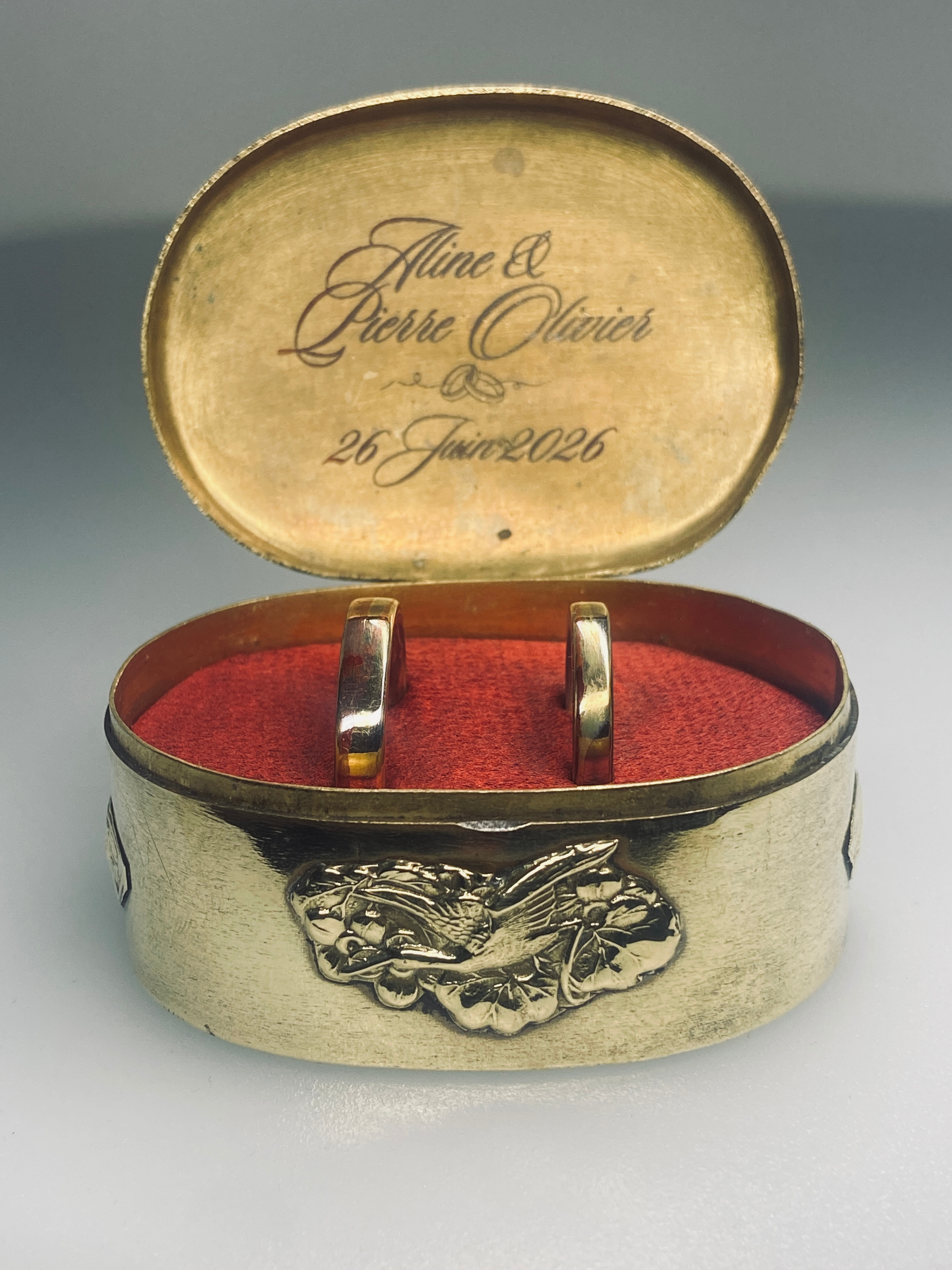 Une boîte en métal dorée contenant deux anneaux de mariage, avec une inscription gravée à l'intérieur du couvercle indiquant les noms 'Aline et Pierre Olivier' et la date '26 juin 2026'.