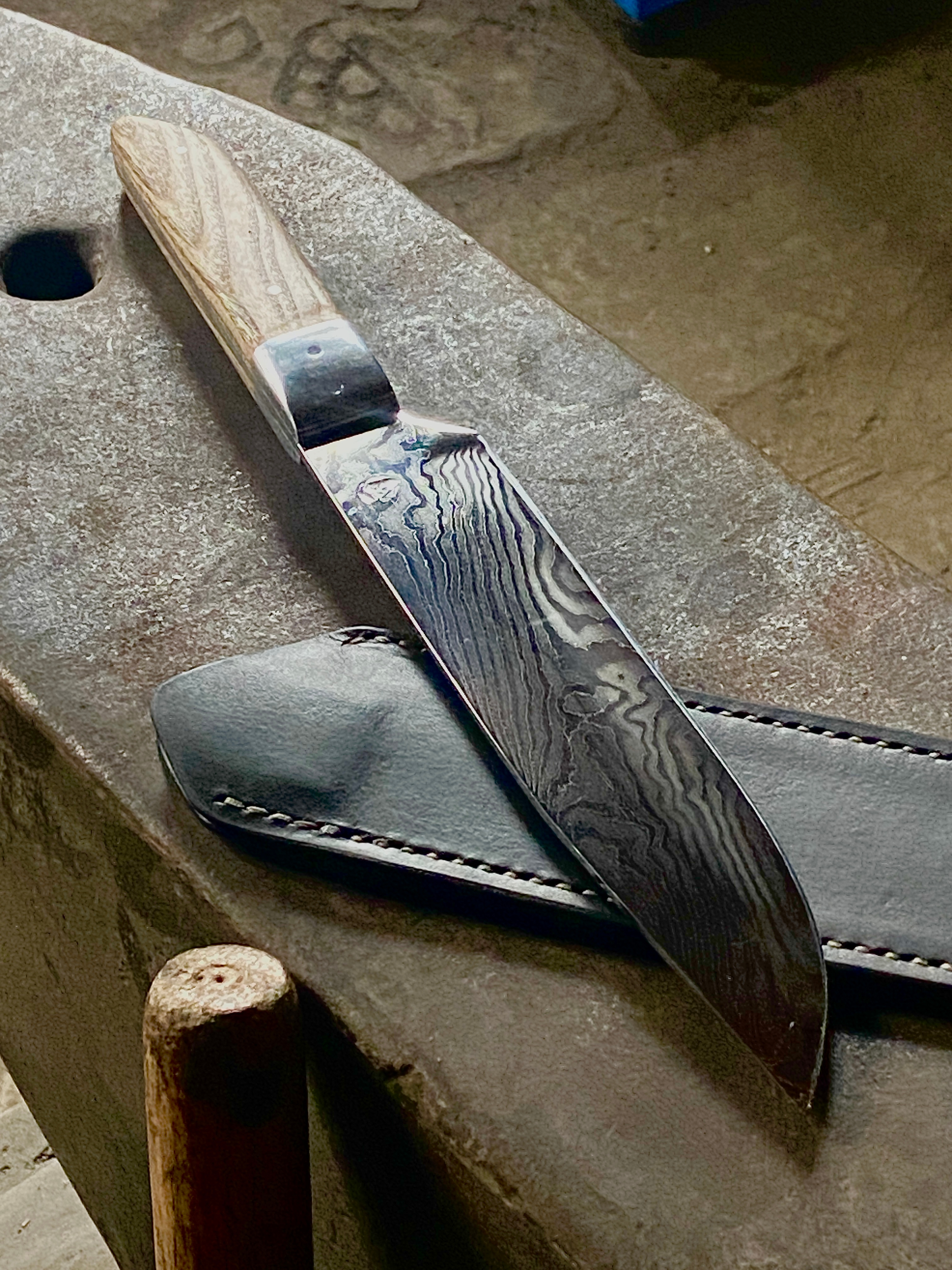 Un couteau de Chef avec une lame Damassée et un manche en bois clair, posé sur un étui en cuir noir, sur une enclume grise.