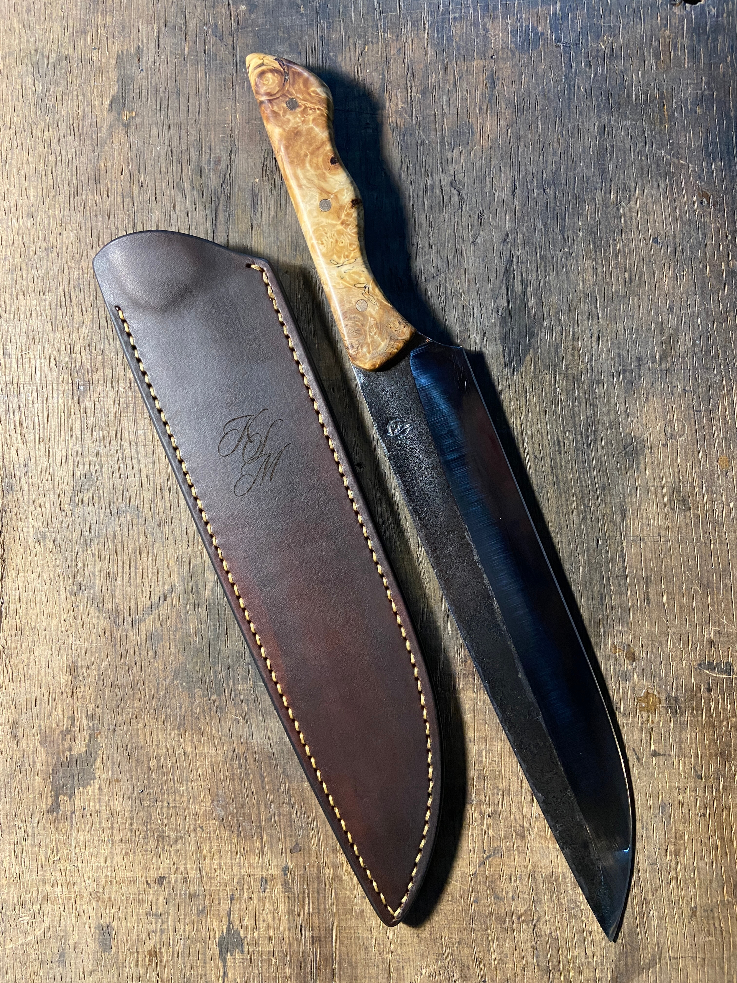 Couteau forgé brut de forge avec manche en bois clair à côté de son étui en cuir brun foncé sur fond de bois de chêne.