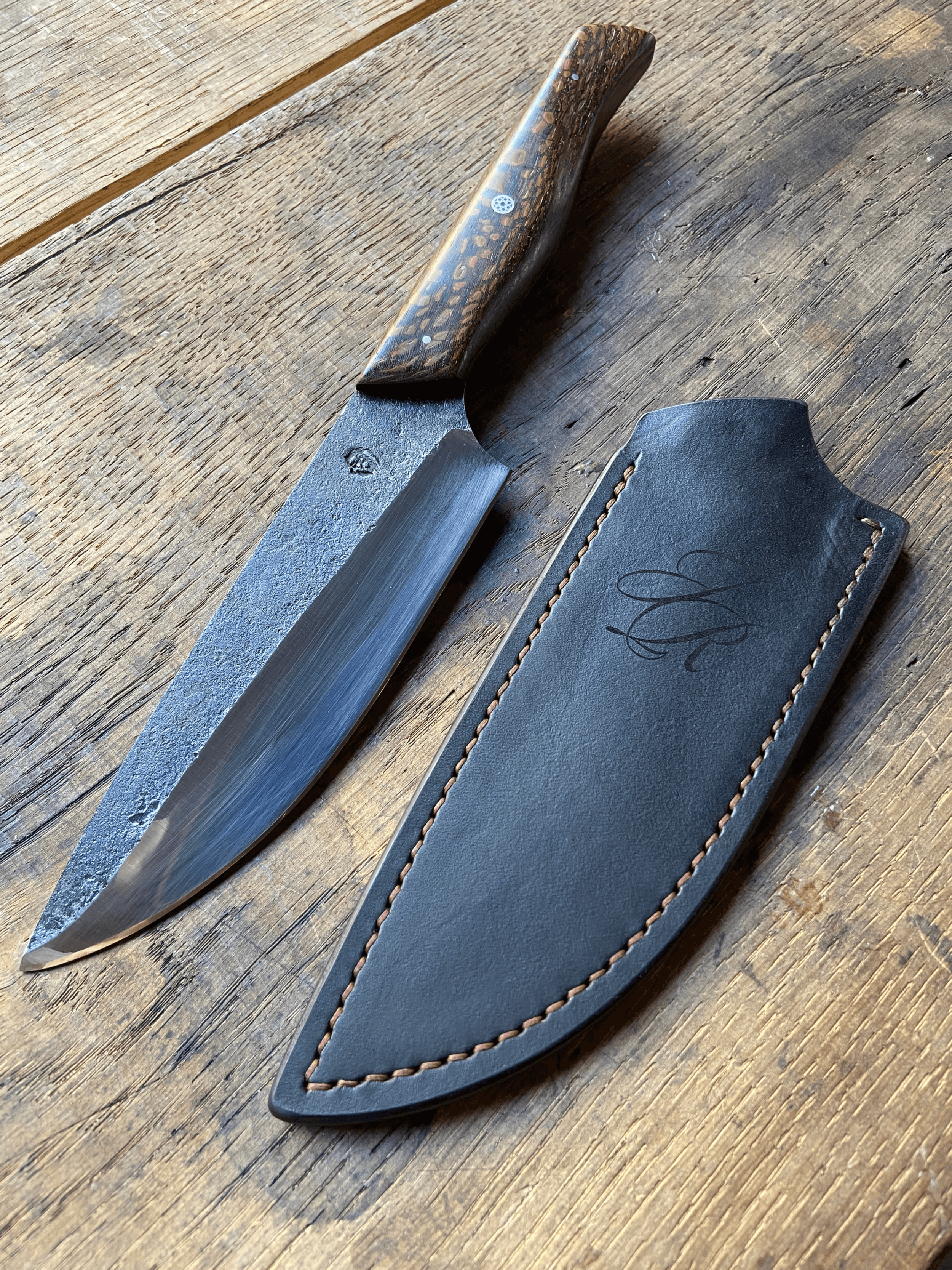 Couteau droit forgé brut de forge avec un manche en bois marron à côté de son étui en cuir marron sur fond de bois de chêne.