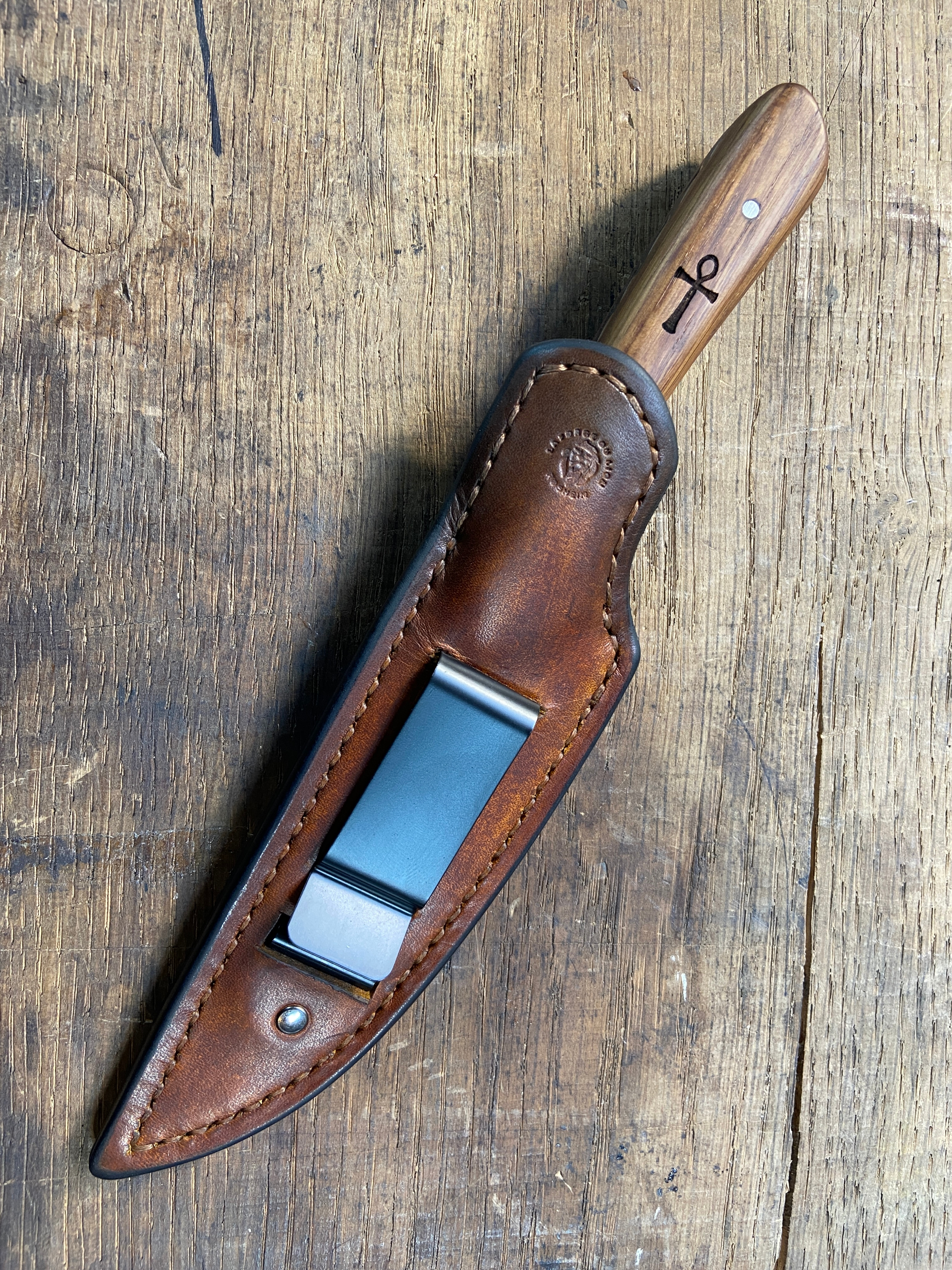 Couteau avec manche en bois brun dans son étui en cuir brun avec du métal noir sur fond de bois.