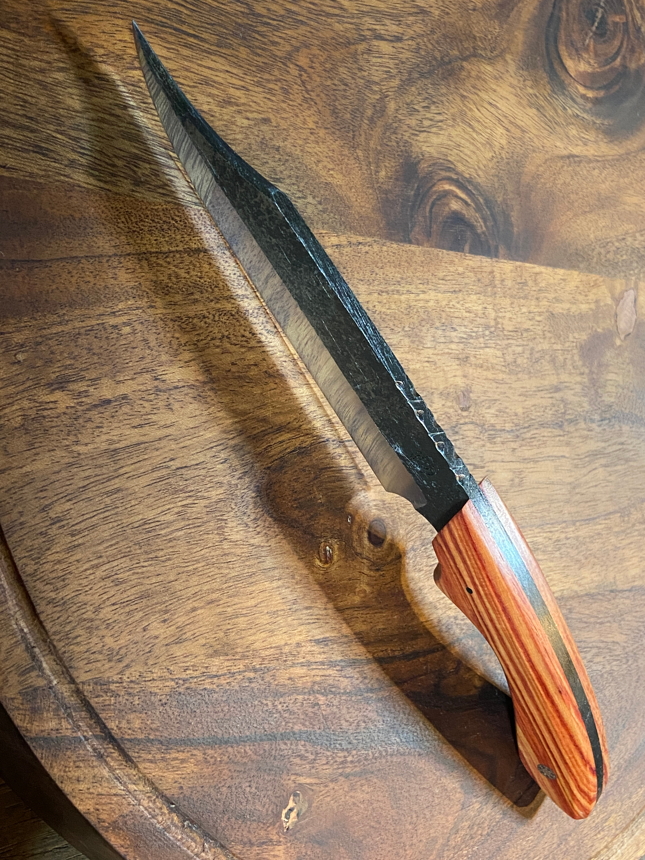 Couteau forgé brut de forge avec manche en bois rouge et blanc sur fond de bois marron.