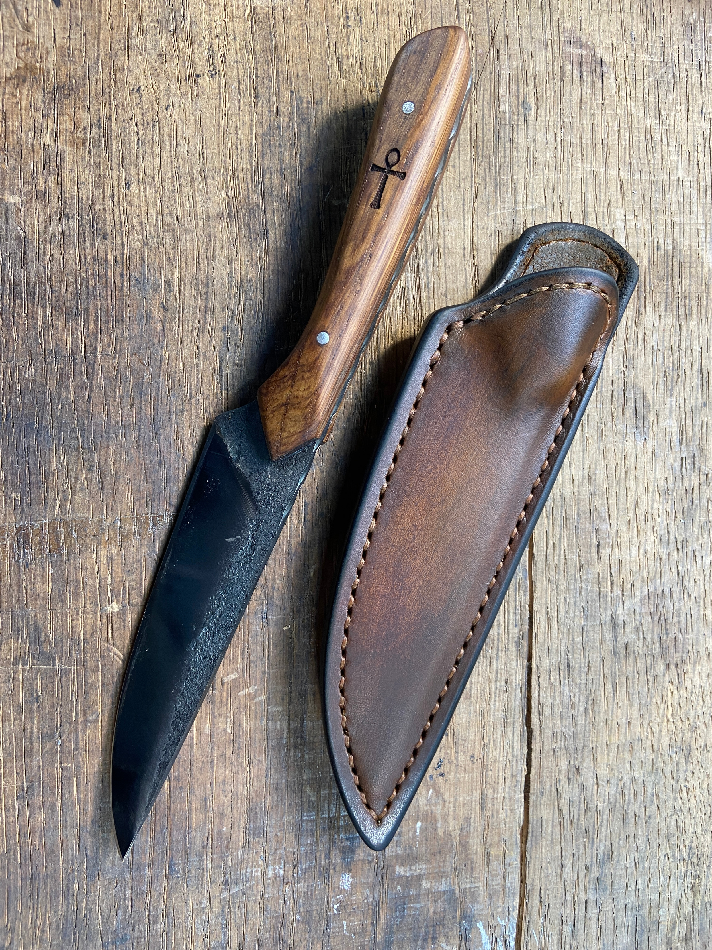 Couteau forgé brut de forge avec manche en bois brun à côte de son étui en cuir brun sur fond de bois.