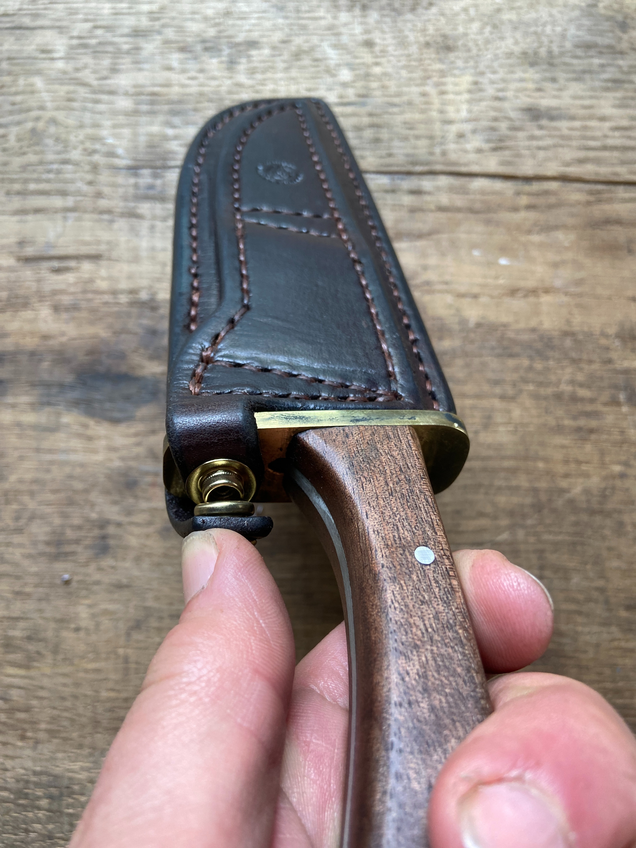 Couteau de chasse avec manche en bois dans son étui en cuir foncé sur fond de bois de chêne clair.