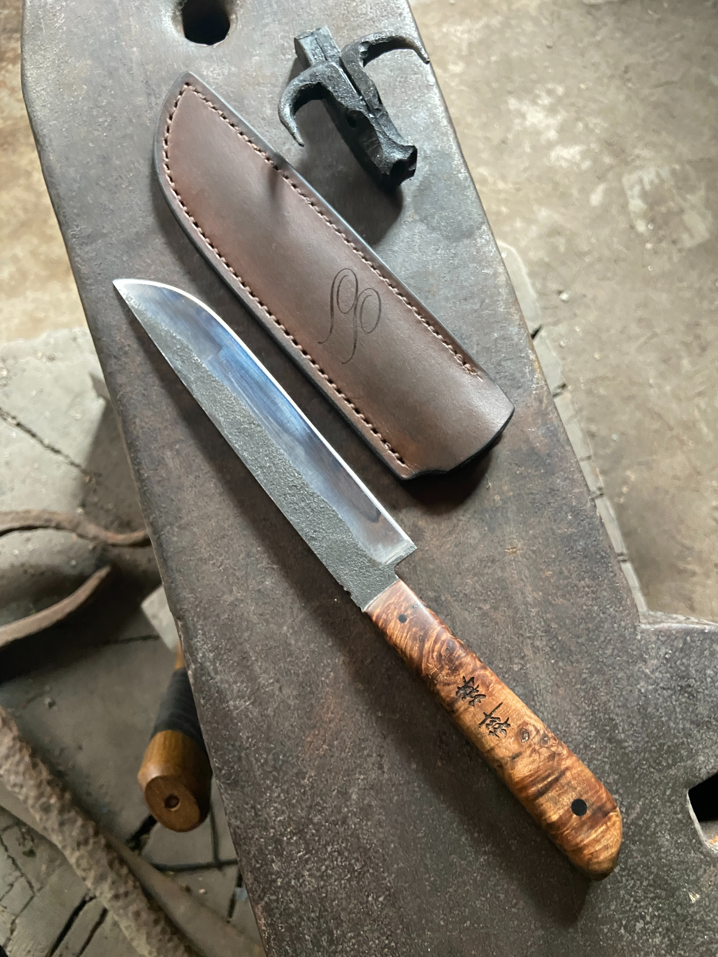 Couteau droit de cuisine forgé brut de forge avec manche en bois stabilisé marron et signes japonais gravés sur le manche sur fond d'enclume grise.