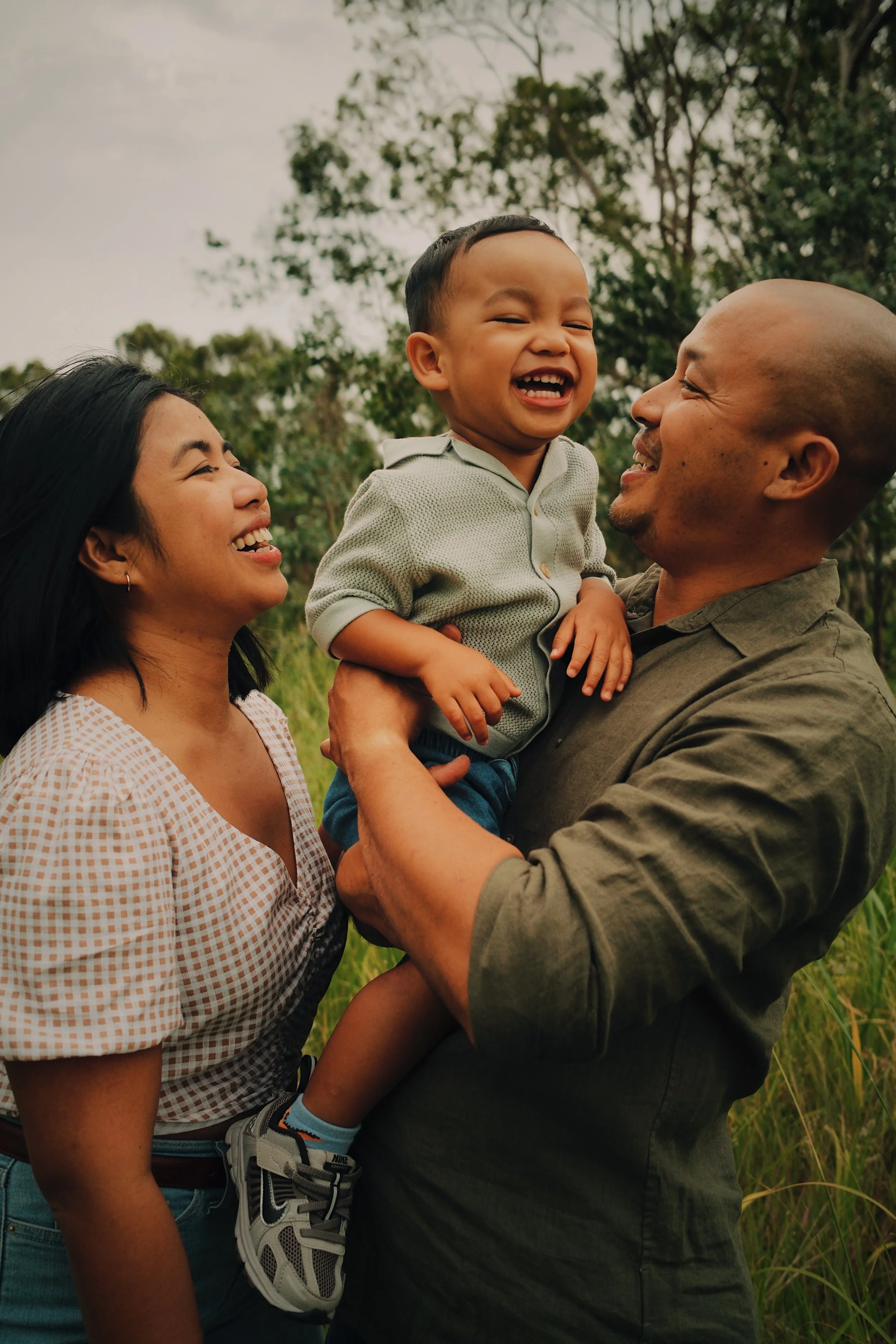 A happy family outdoors with the father holding a young boy and the mother smiling at them, surrounded by trees and greenery.