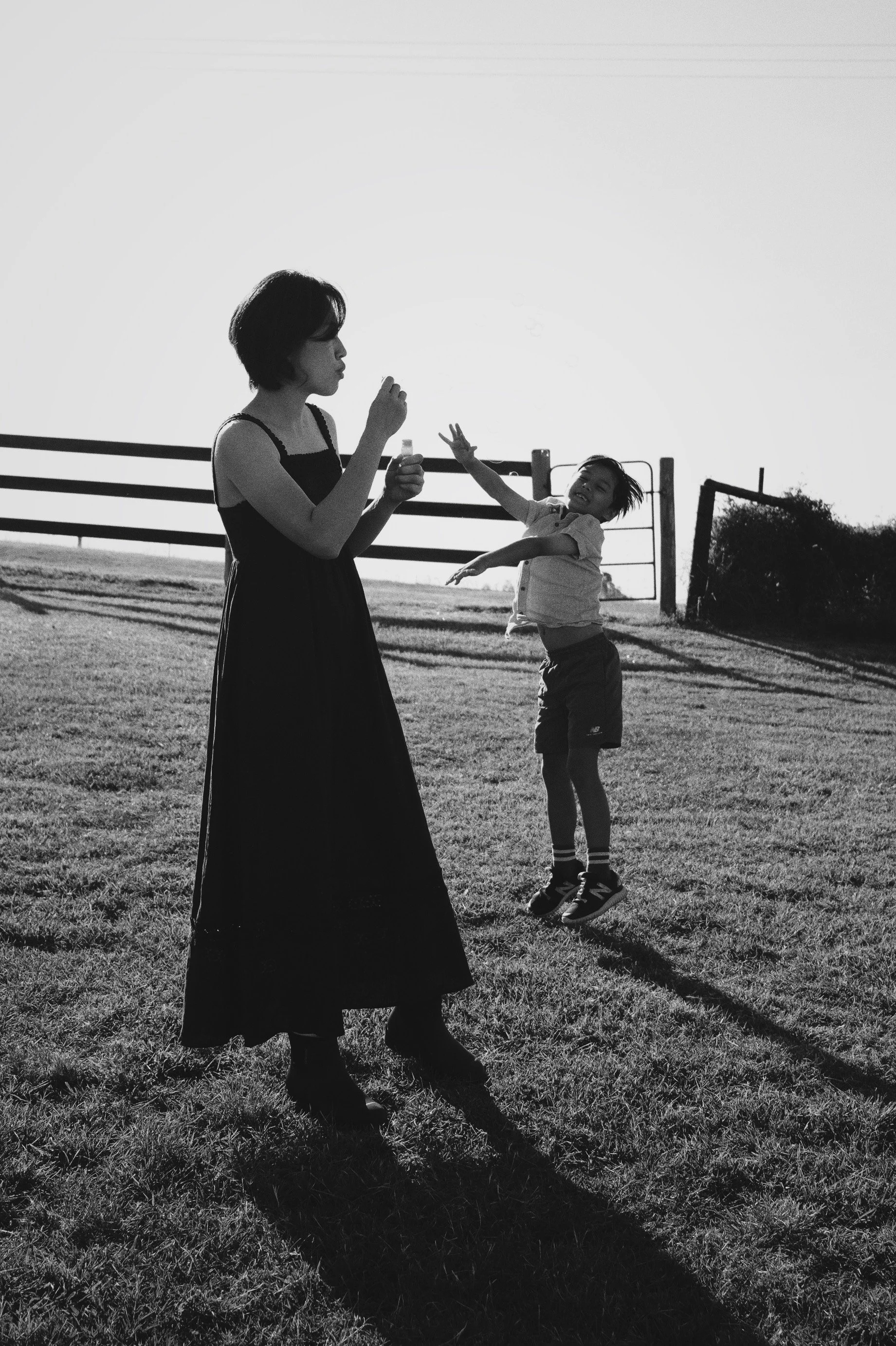 A woman and a young boy outdoors at sunset, with the boy jumping and reaching out towards the woman, who is holding a small object, on grassy field with fence and trees in the background.