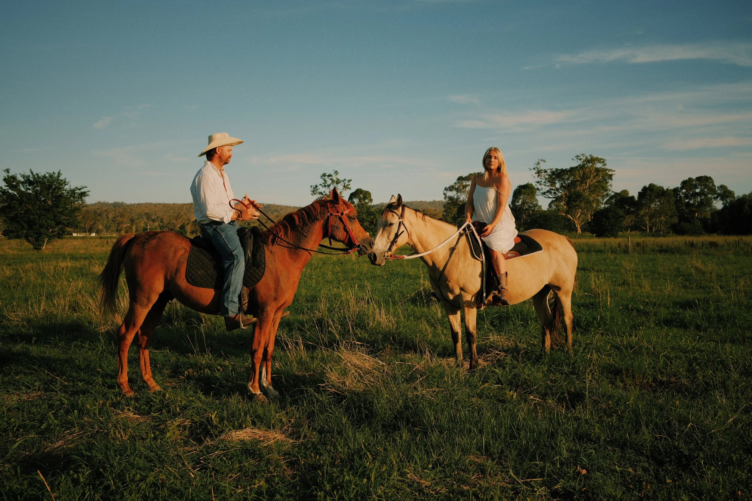 A man and woman riding horses in a grassy field at sunset, with trees and hills in the background.