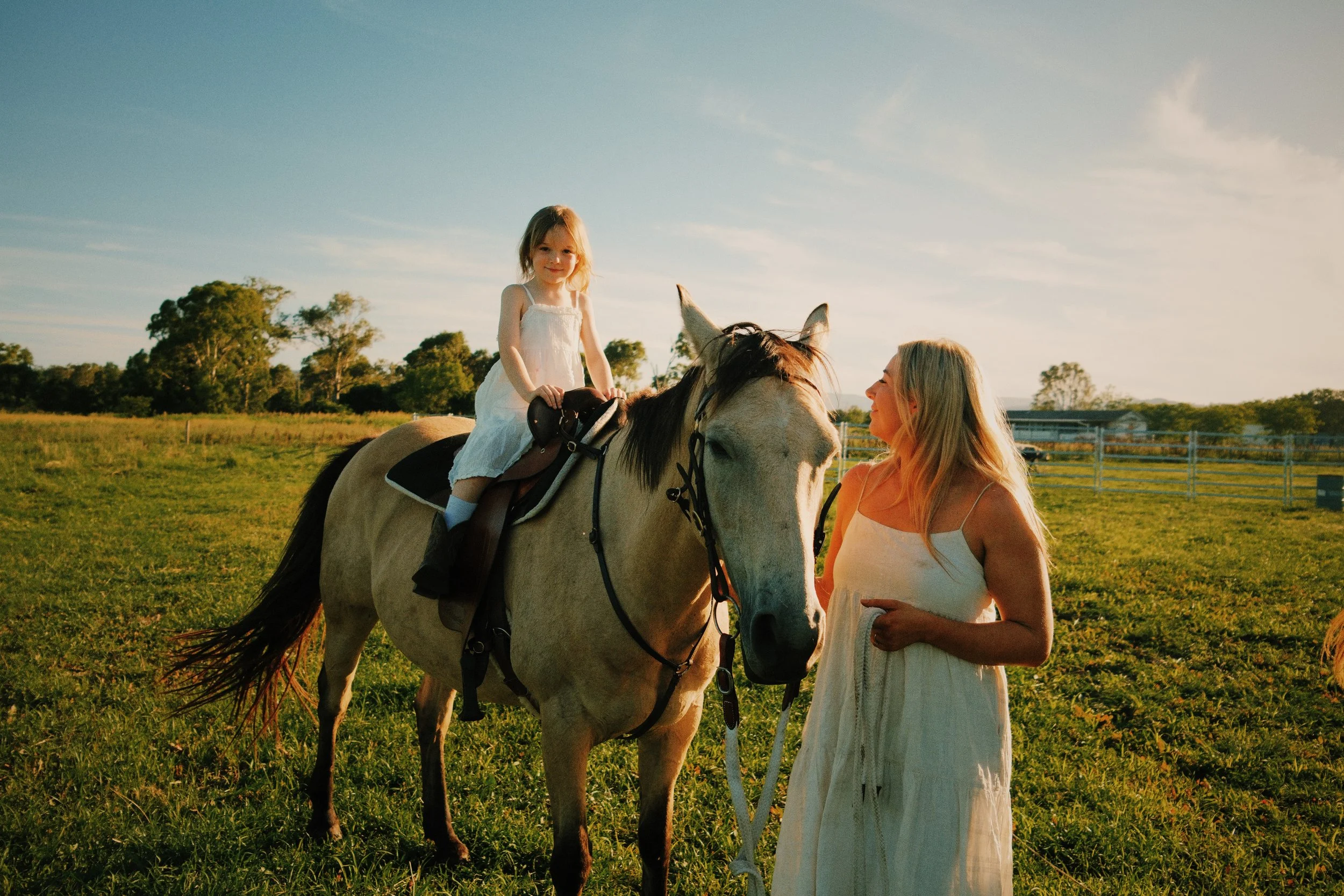A young girl in a white dress riding a light-colored horse in a grassy field, with a woman in a white dress standing nearby, holding the horse's reins, both smiling and looking at each other during sunset.
