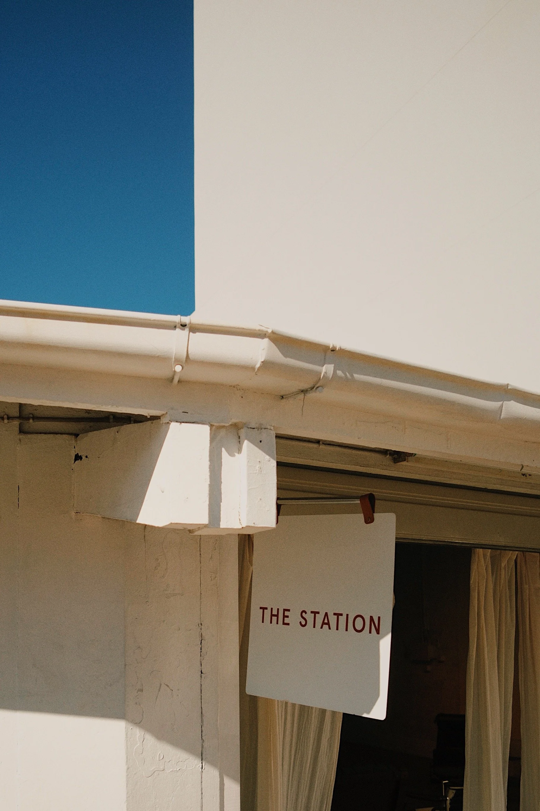 A white exterior wall with a sign that reads 'The Station' hanging near a door with beige curtains. Part of the blue sky is visible above.