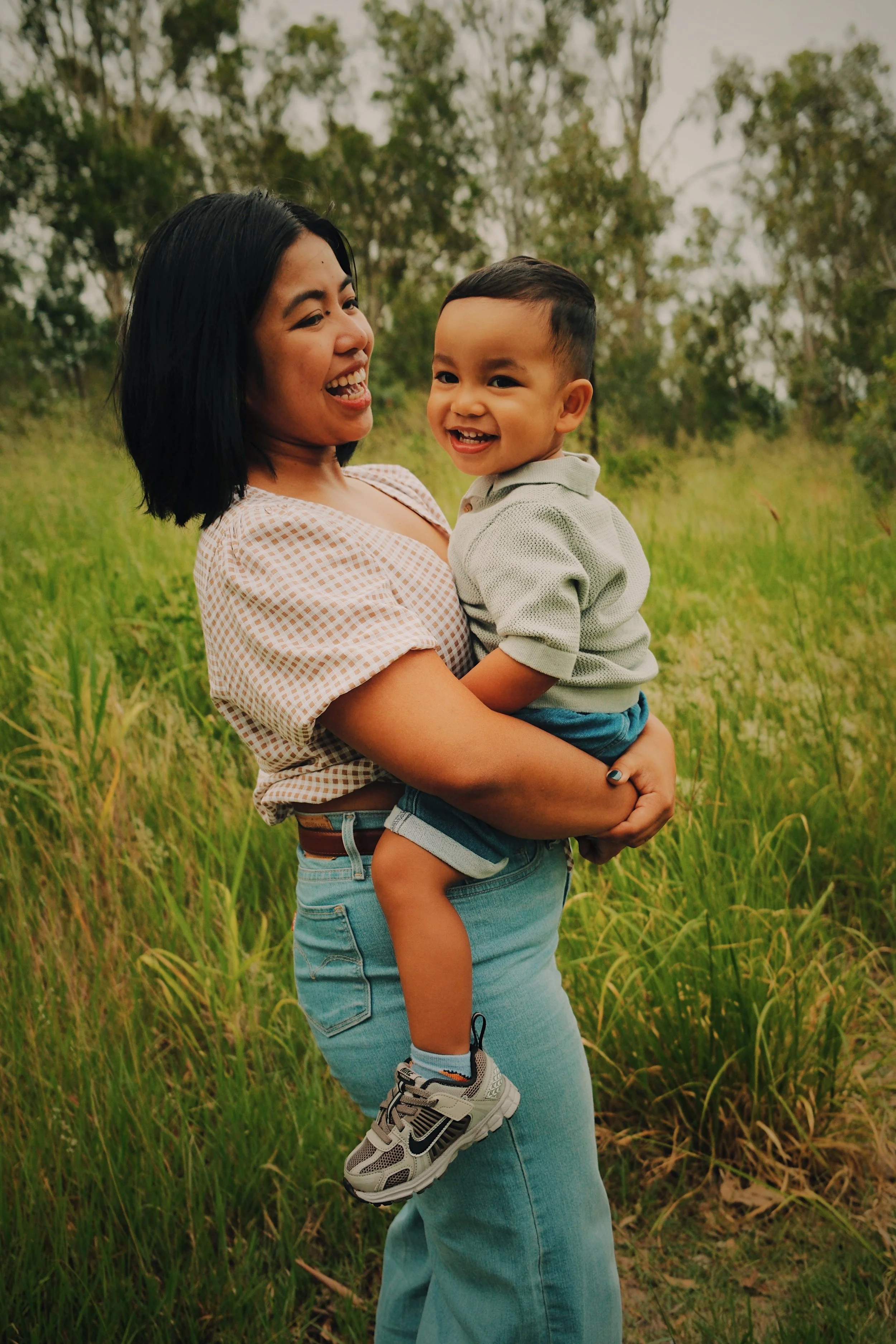 A woman holding a smiling young boy in a grassy outdoor area with trees in the background.
