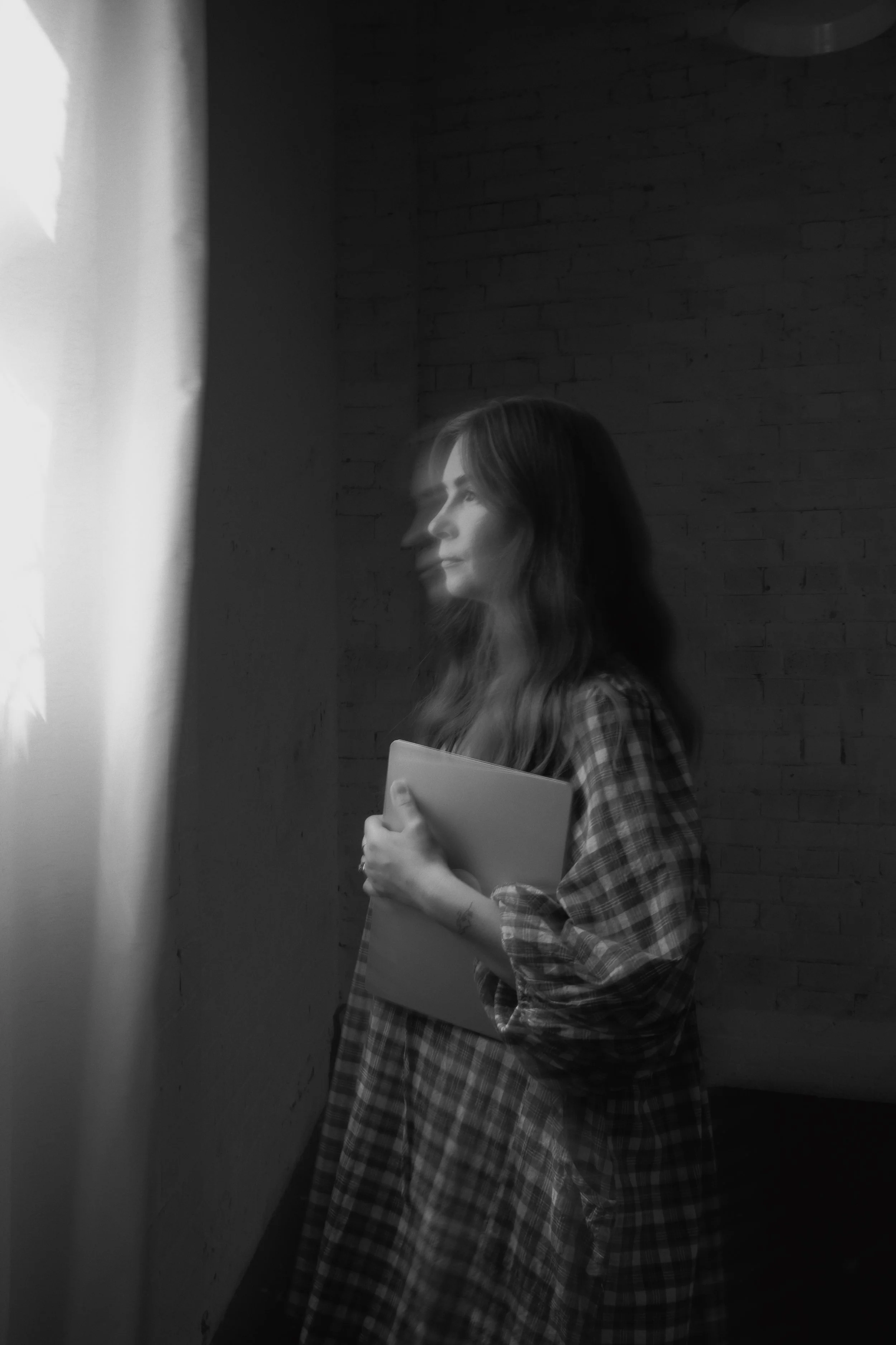 A black and white photo of a woman standing near a window, holding a closed laptop, gazing outside with a thoughtful expression.