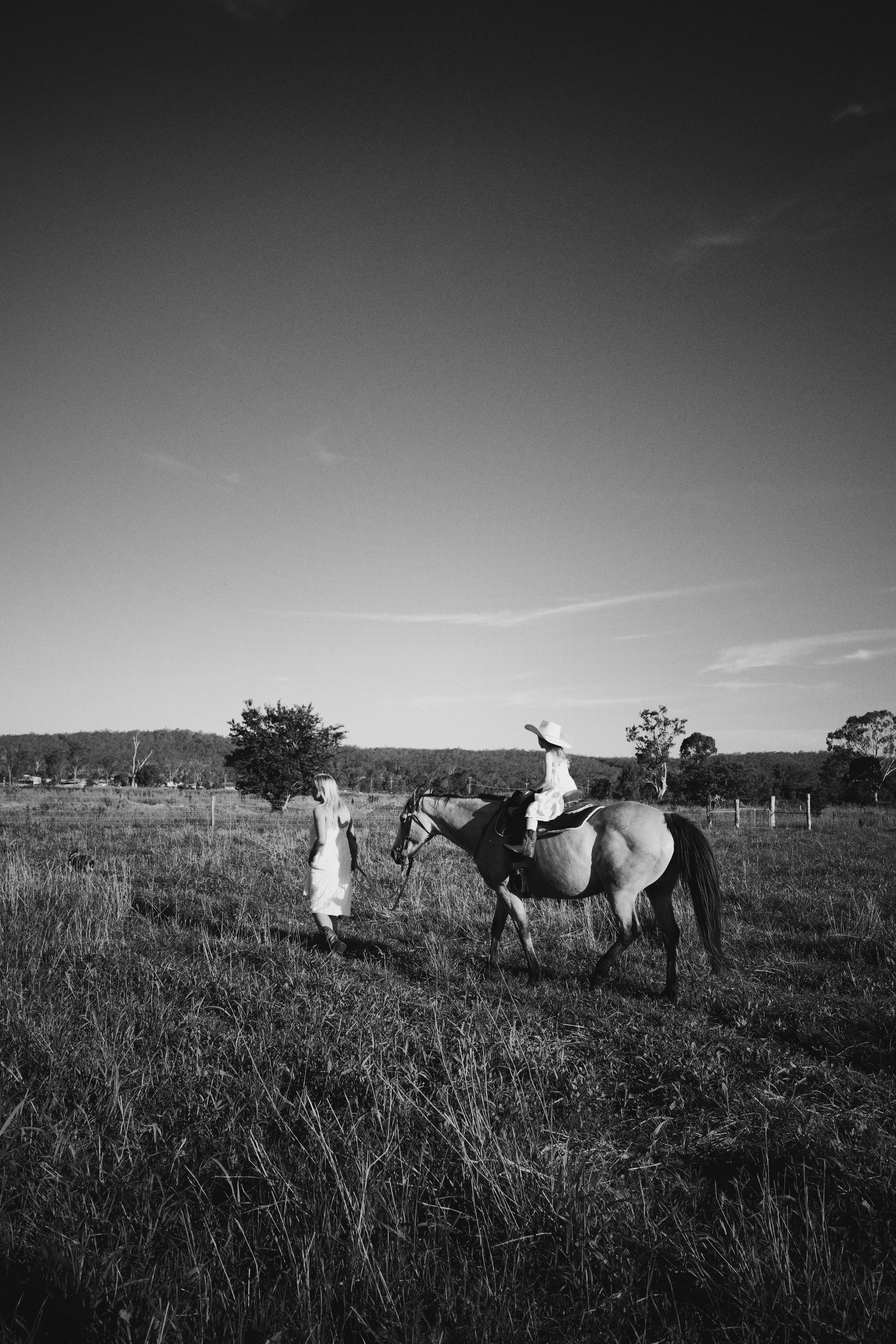 A black and white photo of two women on a farm or countryside, one walking ahead and the other riding a horse, under an open sky with sparse clouds.