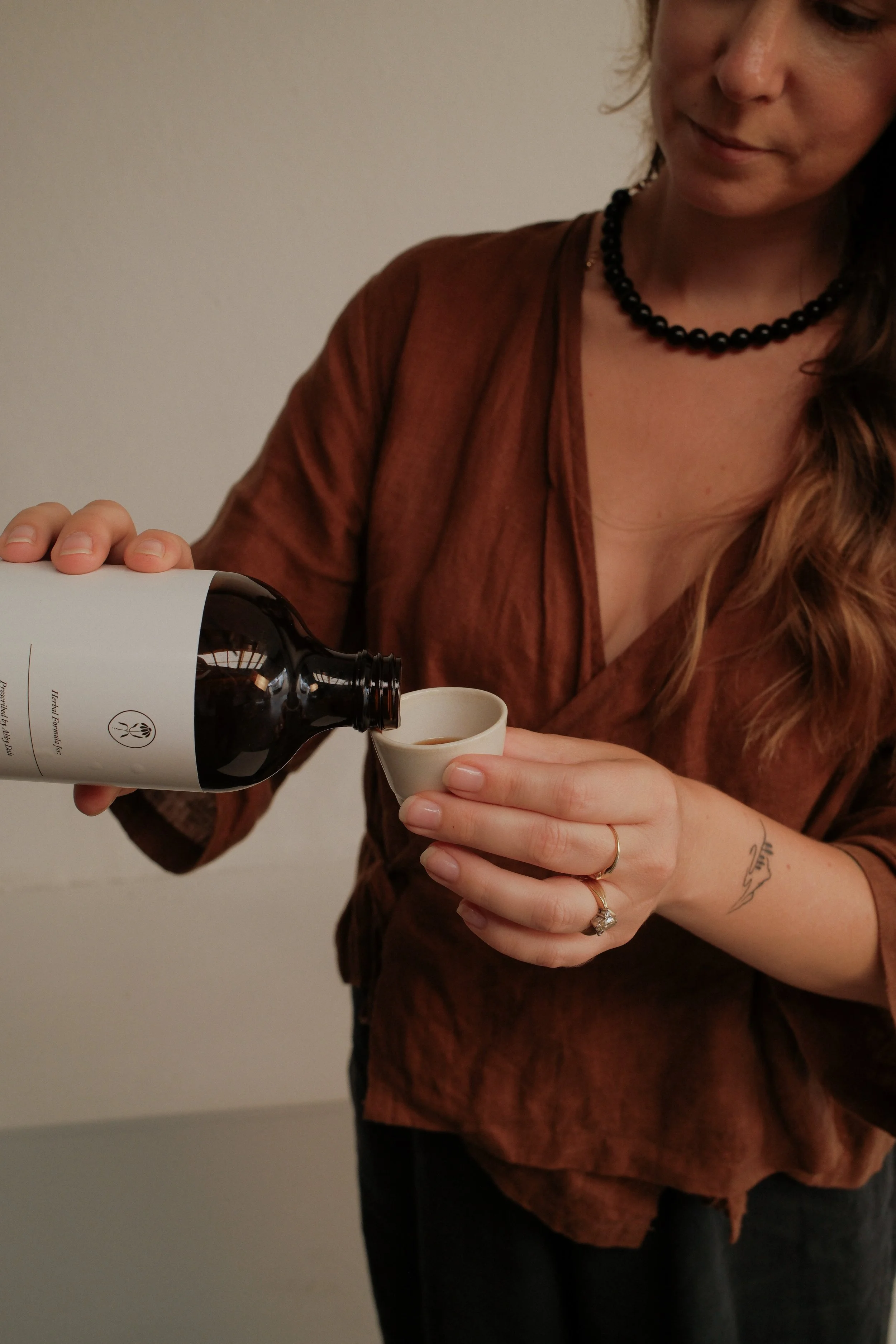 A woman pours a dark beverage from a glass bottle into a small white cup.