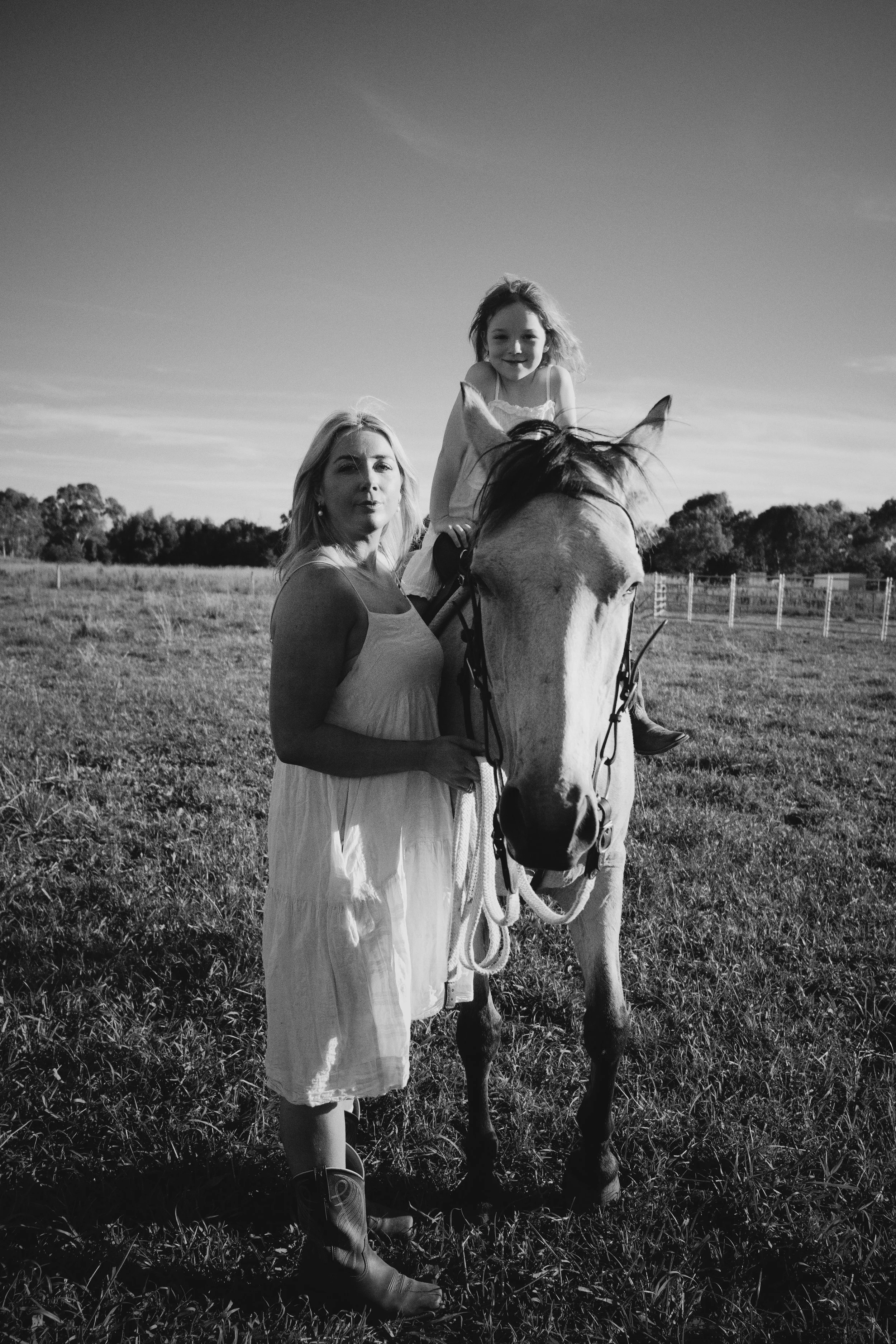 A woman and a young girl with curly hair riding a horse in a grassy field on a sunny day, black and white photo.
