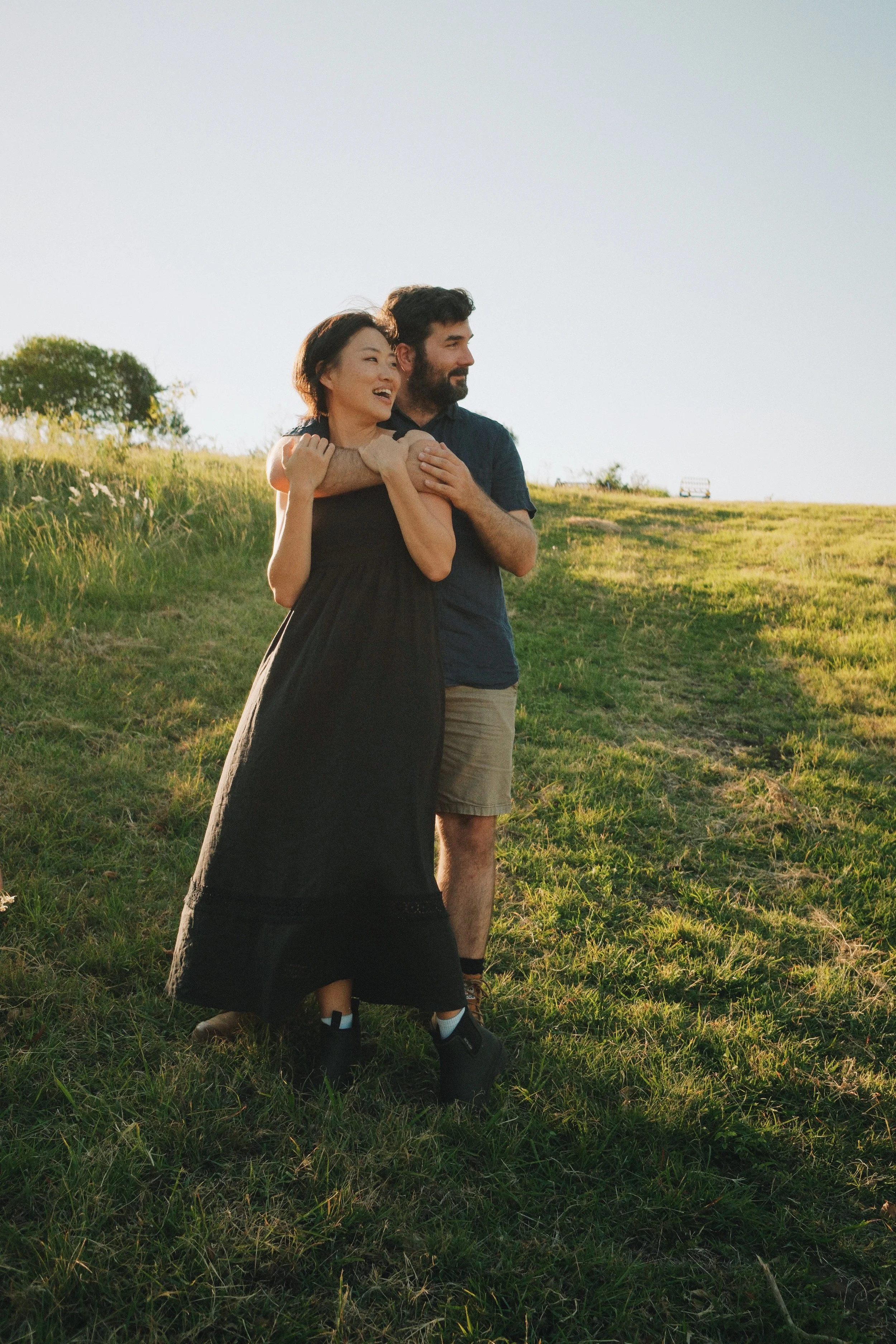 A couple standing on a grassy field during sunset, embracing each other and smiling.