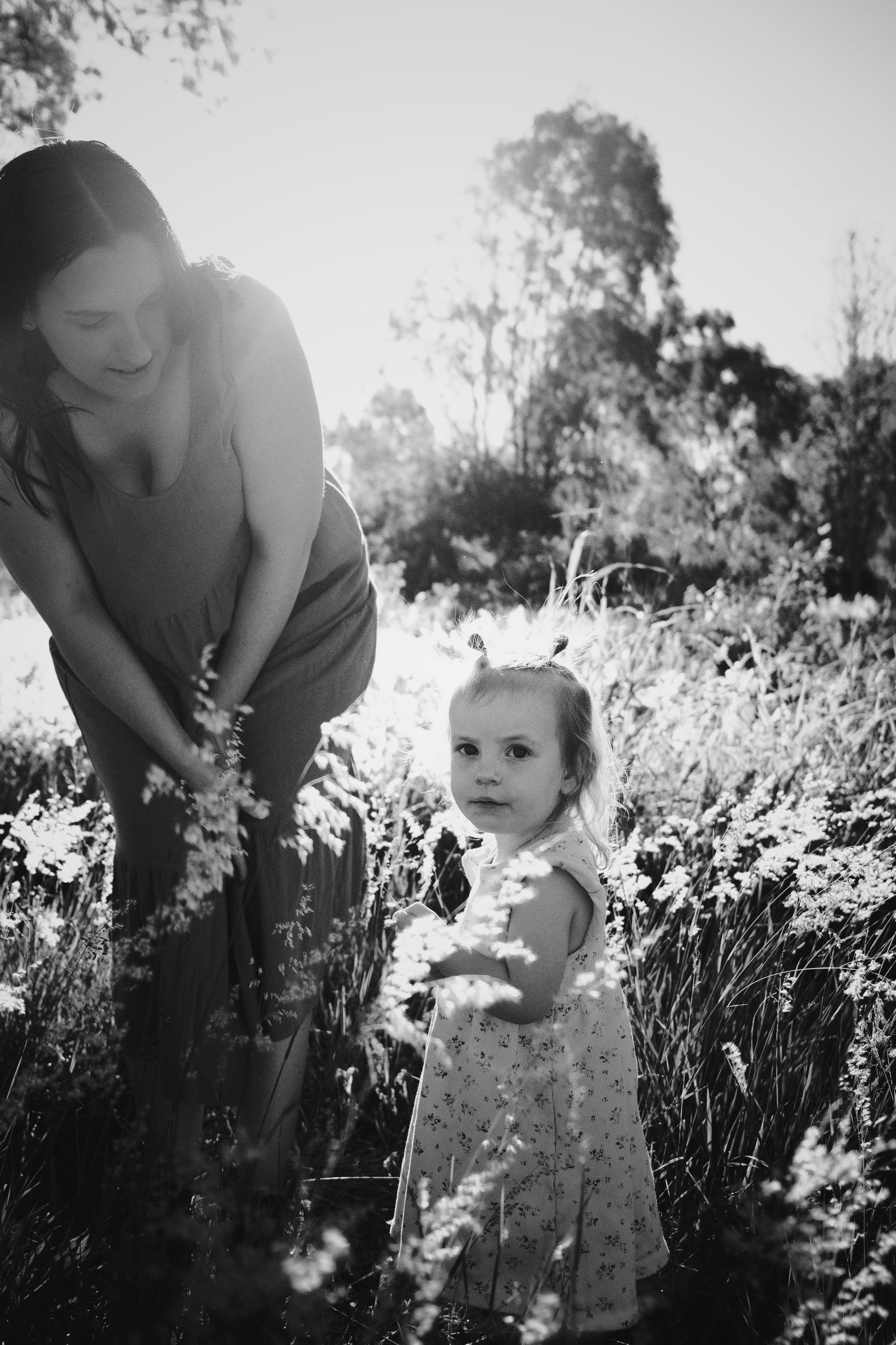 A woman and a young girl standing in a field of tall grass and wildflowers, with trees in the background and sunlight shining from behind.