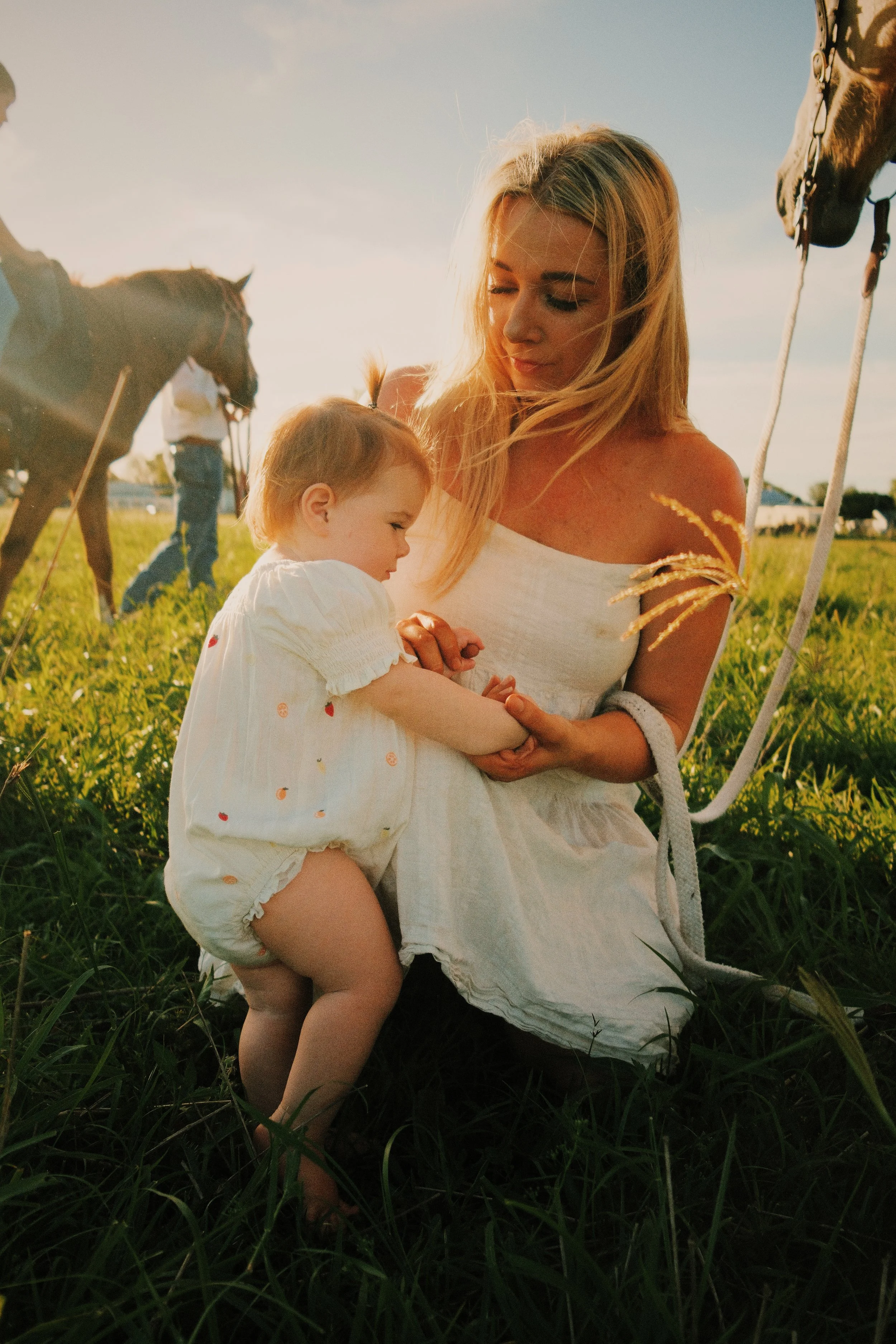 A woman with long blonde hair sitting in a grassy field during sunset, holding a young child with short blonde hair, and two horses in the background, one on each side.
