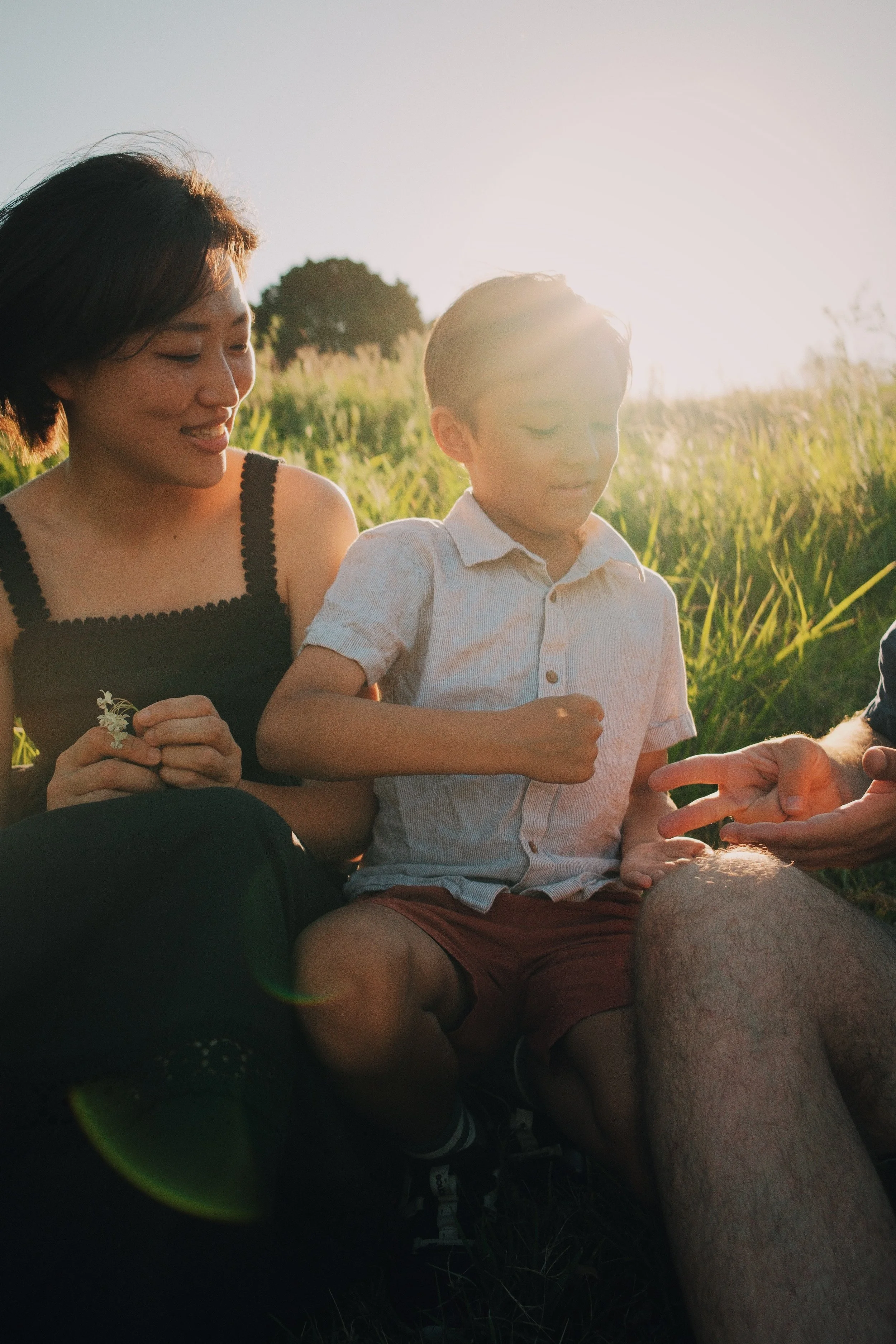 A woman and a young boy sitting outdoors in a grassy field during sunset, with a hand showing a flower.
