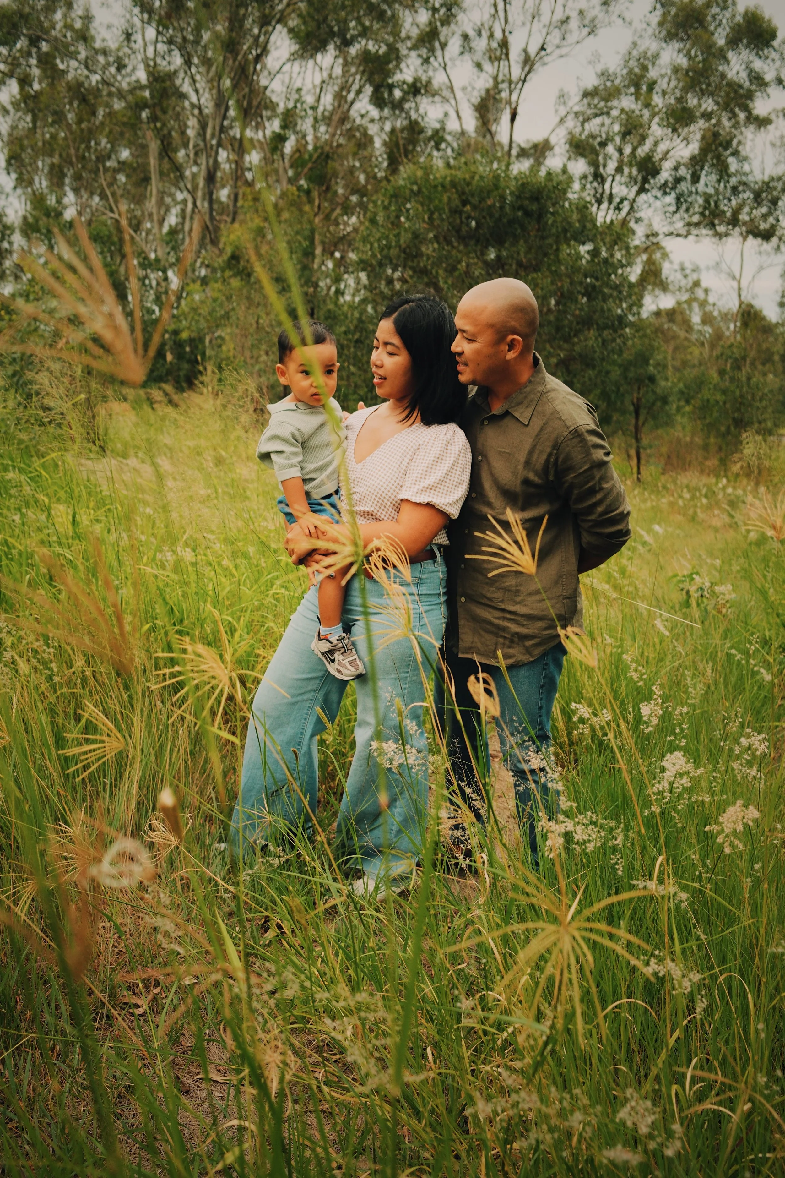 A family of three standing in a grassy field with trees in the background. The mother holds a young boy, and the father stands close, all engaging with each other.
