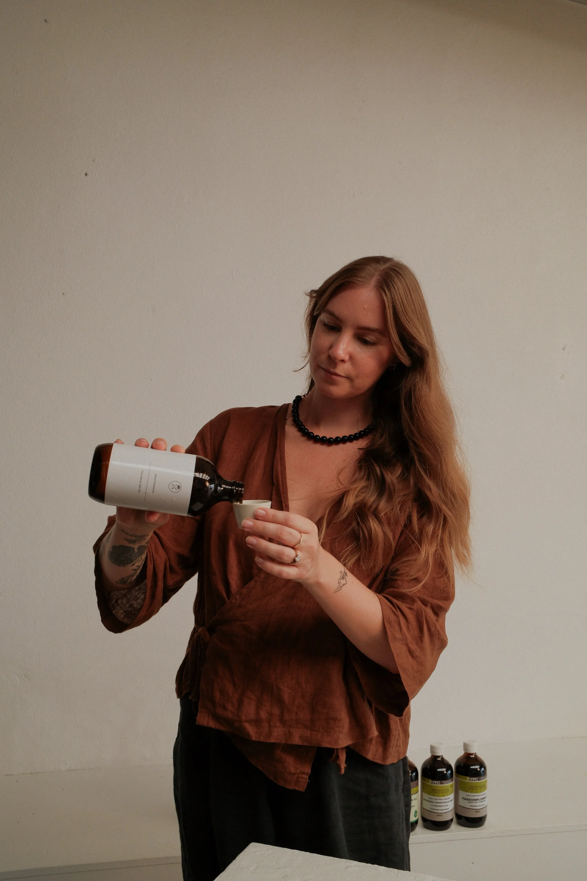 A woman with long red hair, wearing a brown blouse and a black bead necklace, pours a liquid from a dark bottle into a small white cup.