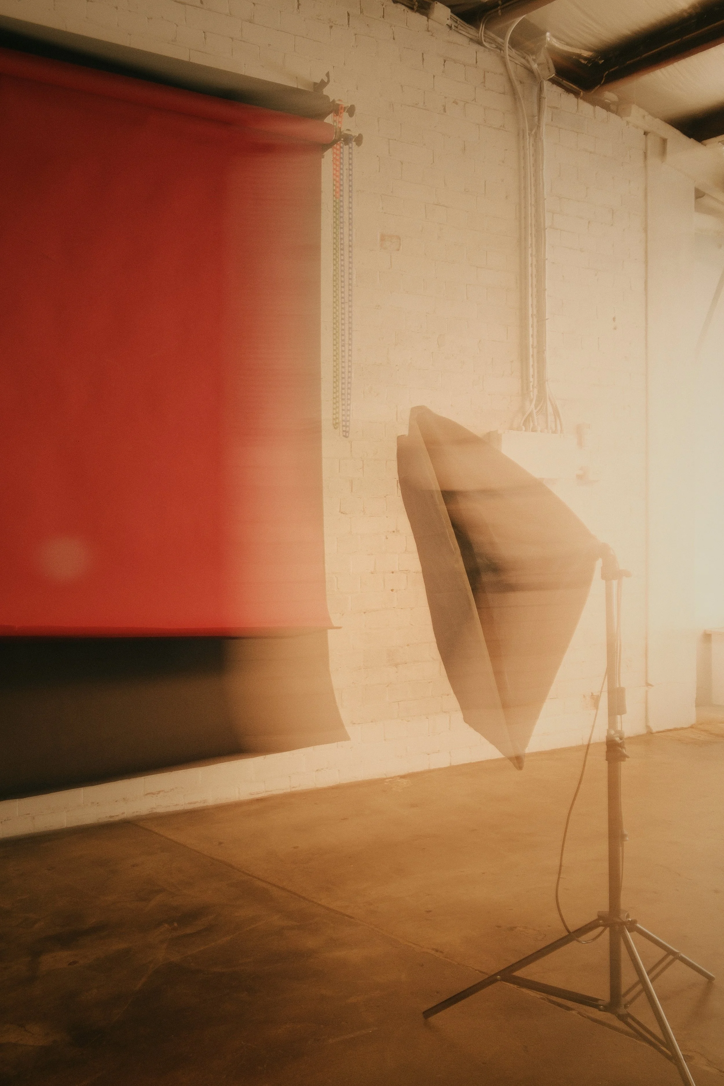Photo of a photography studio setup with a softbox light, a reflector, and a red backdrop against a white brick wall.
