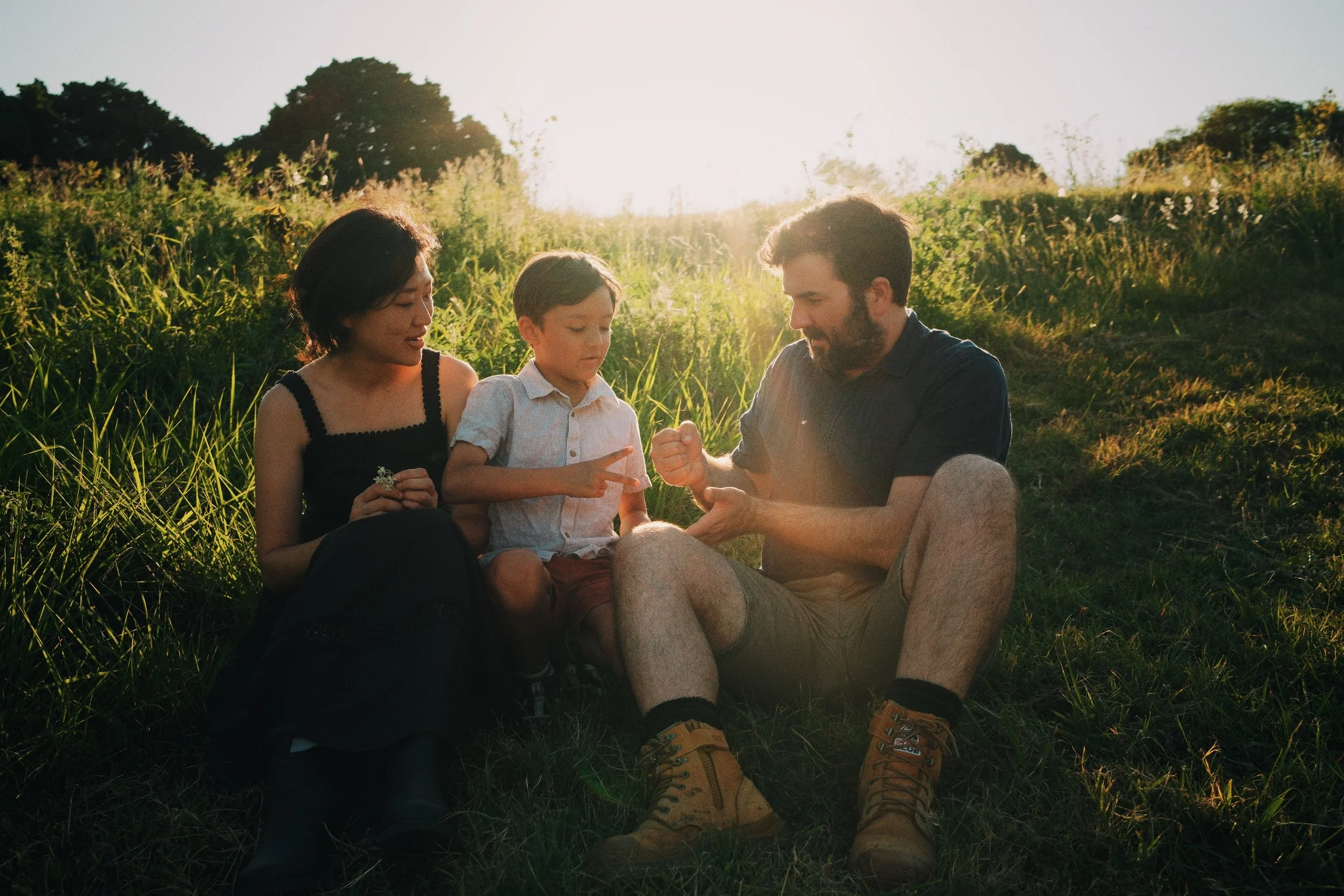 A family of three sitting on grass in a field during sunset, with the father showing something to his son, while the mother looks on.
