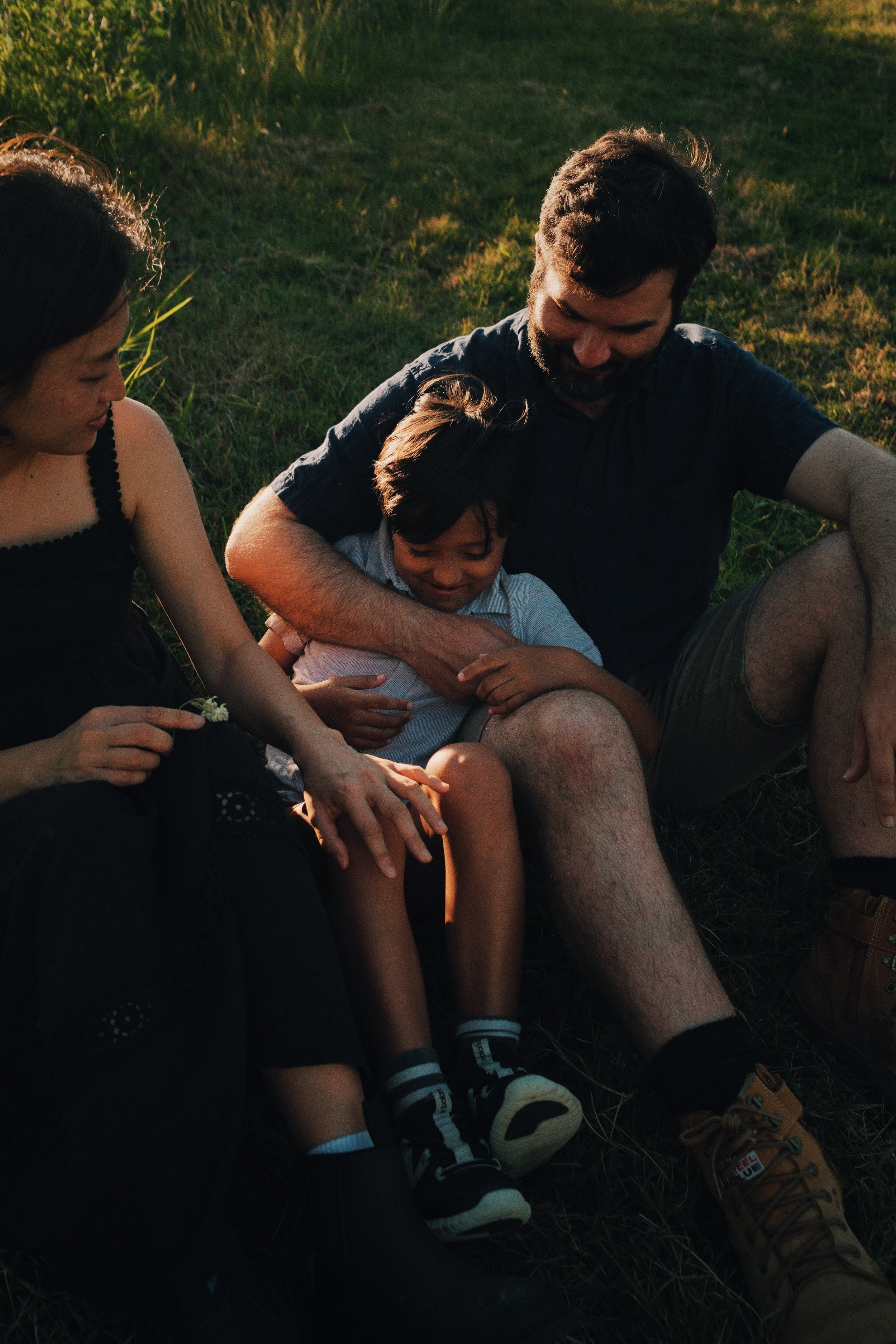 Family sitting on the grass, sharing a joyful moment during sunset, with a woman on the left, a man on the right, and a boy in the middle leaning into the man.
