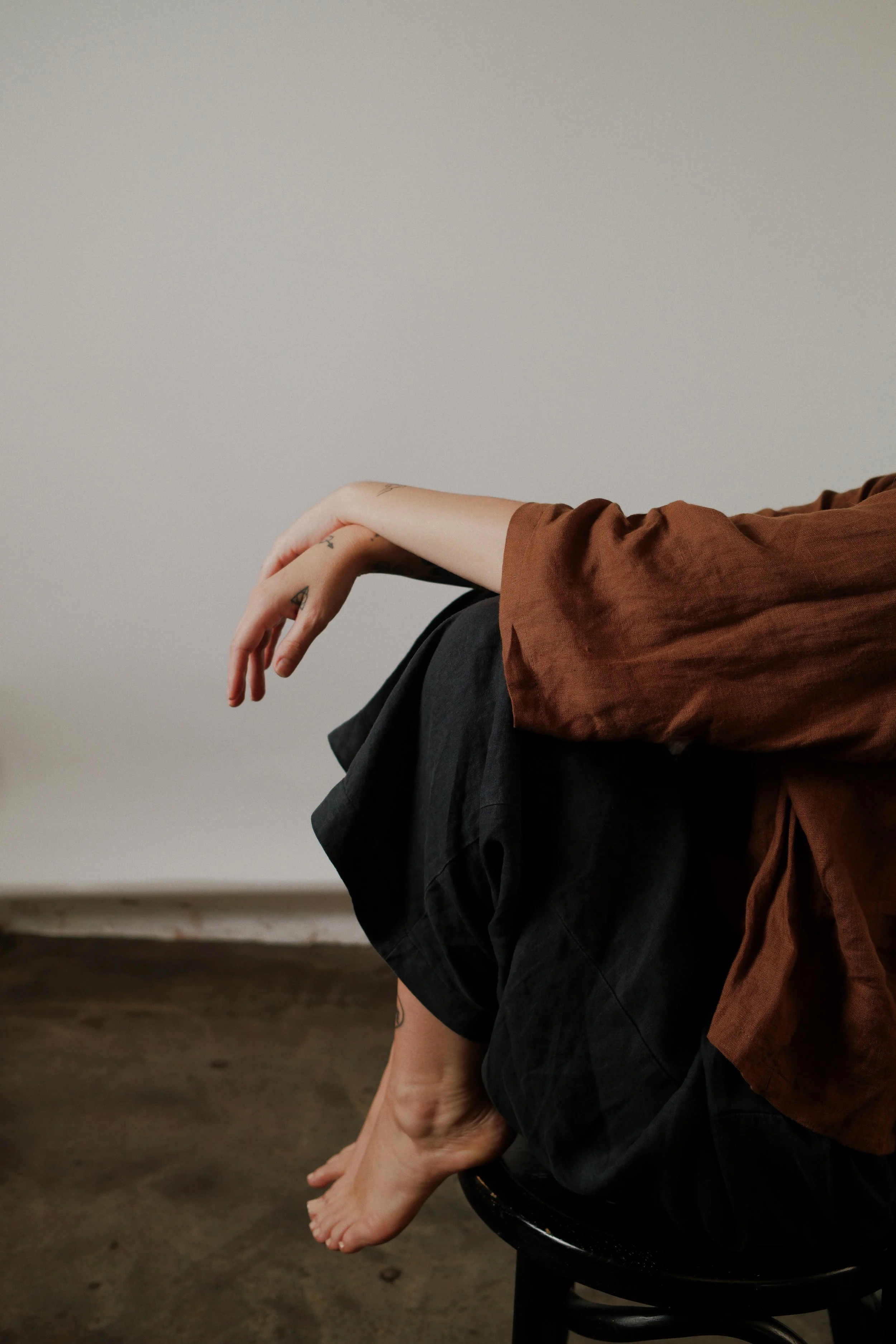 A person sitting on a black stool with their legs and feet hanging over the side, wearing black pants and a brown top, with a bare foot visible. The person's arms are resting on their knees, visible tattoos on their fingers and arm. The background is