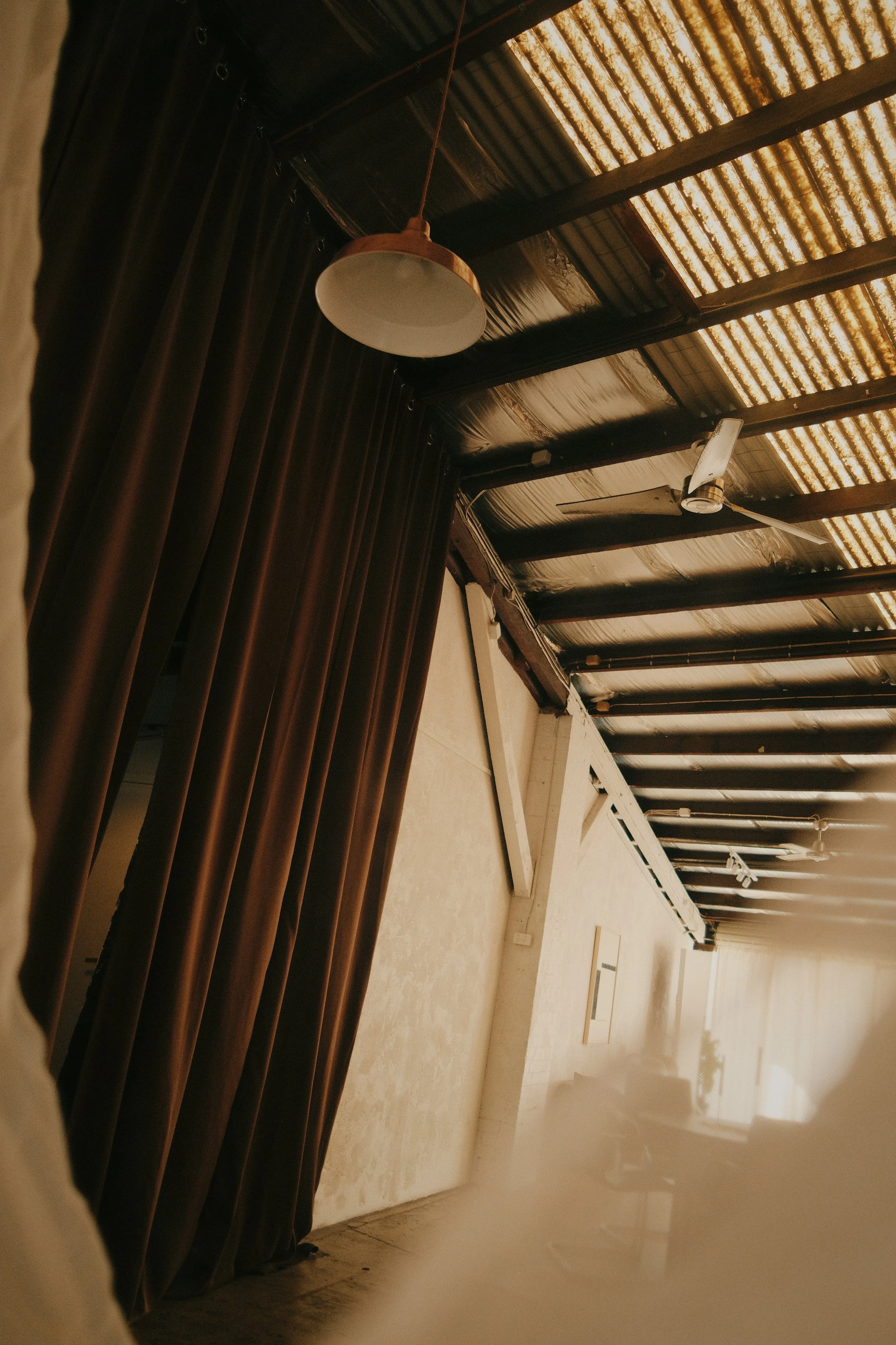 Interior of a room showing a black curtain, a wooden ceiling with exposed beams, a ceiling fan, an overhead light, and a white wall with a mounted white frame.