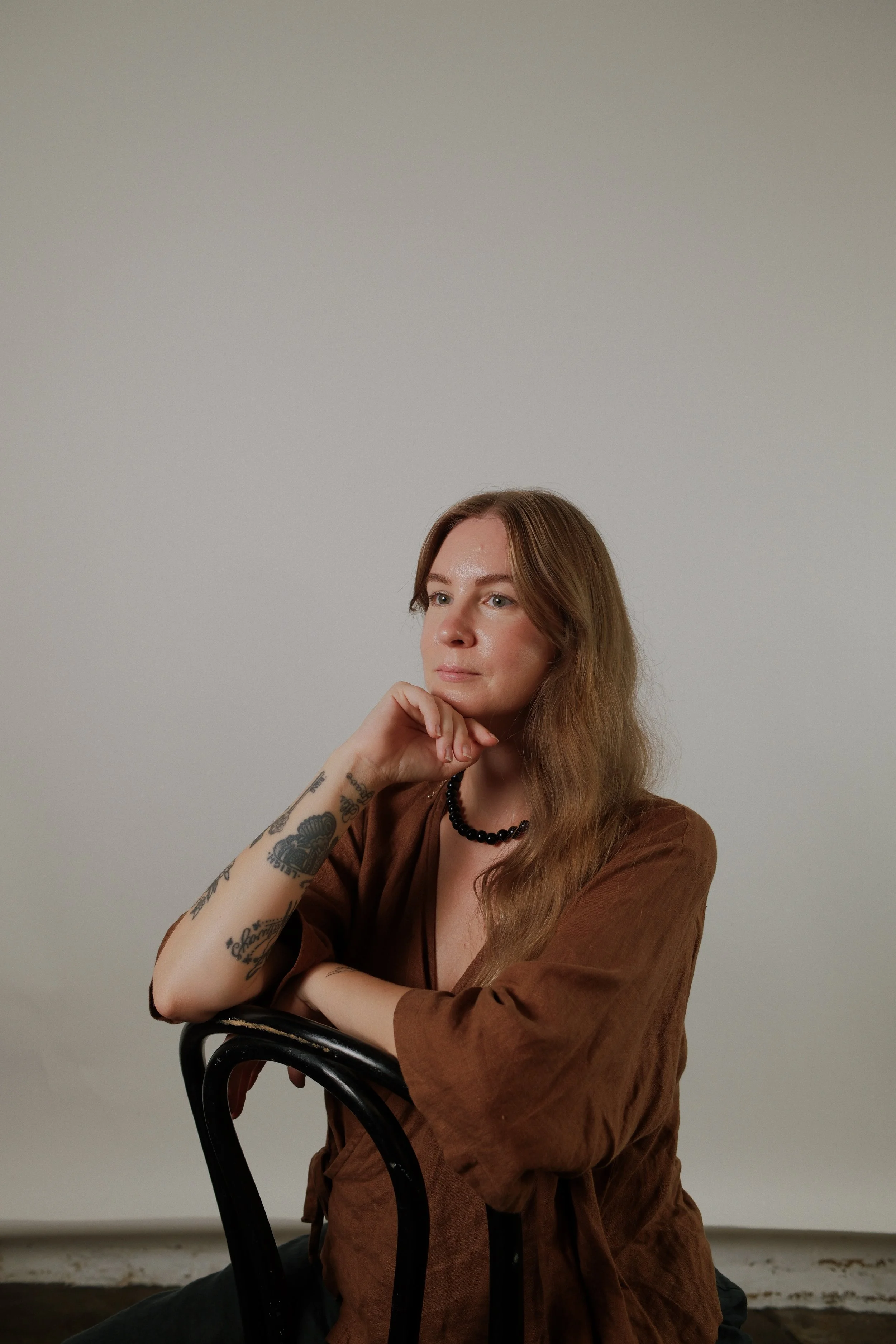 A woman with long, wavy red hair, wearing a brown top, black beaded necklace, and tattoos on her forearm, sitting on a black chair with her arm resting on the backrest, looking thoughtfully to the side against a plain, light gray background.