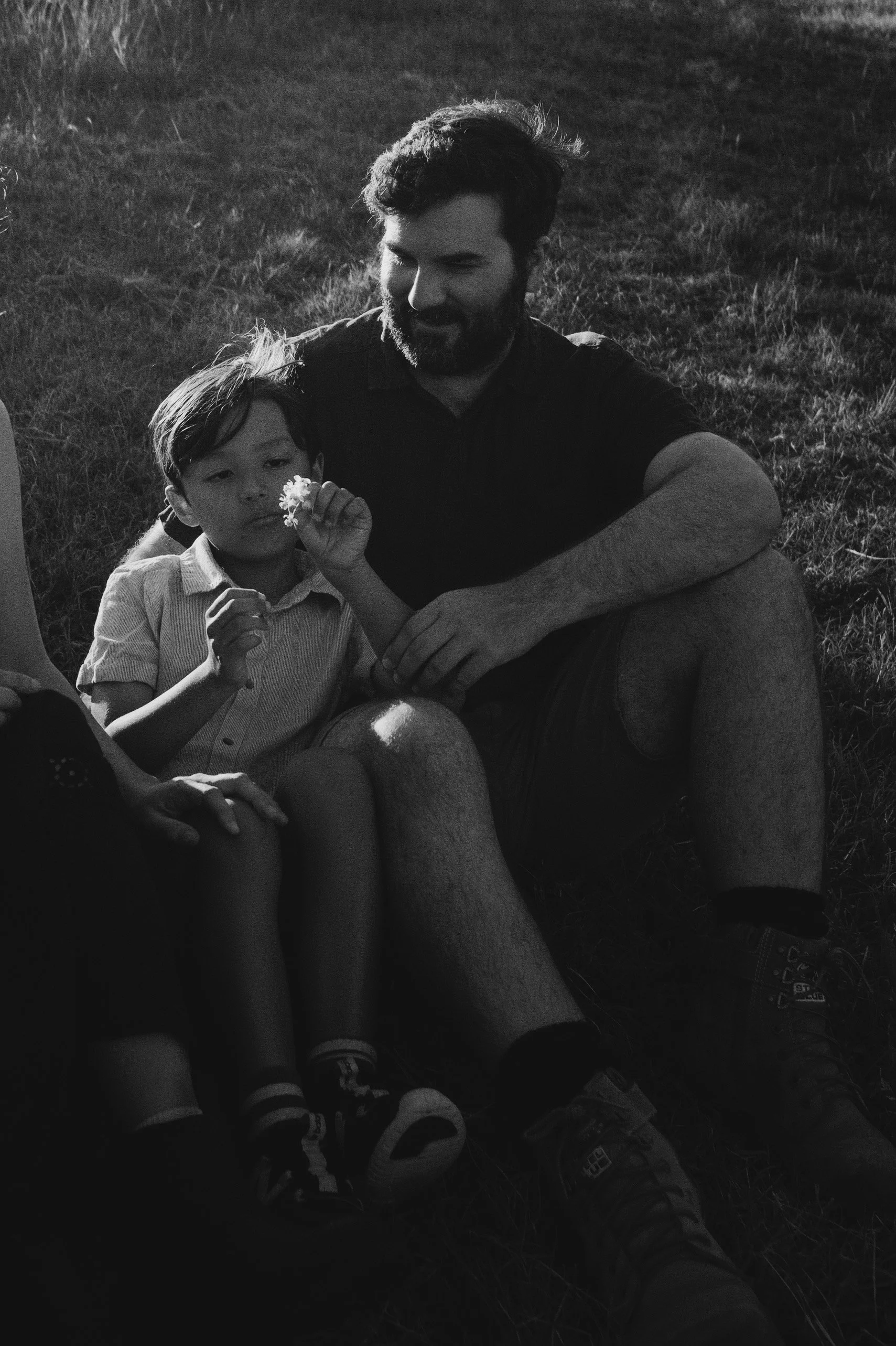 A man and a young boy sitting on the grass outdoors, with the boy holding a flower and looking at the camera, in a black and white image.