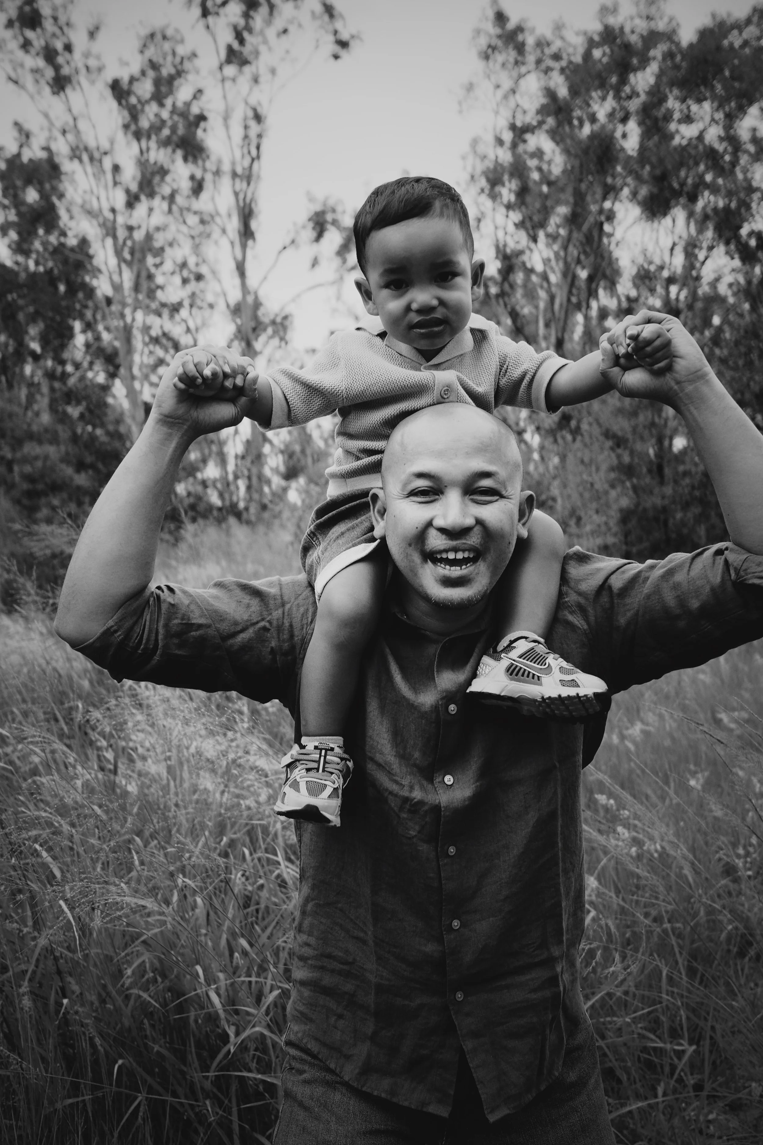 A black and white photo of a man carrying a young boy on his shoulders in an outdoor grassy area with trees in the background. The boy and man are flexing their muscles and smiling.